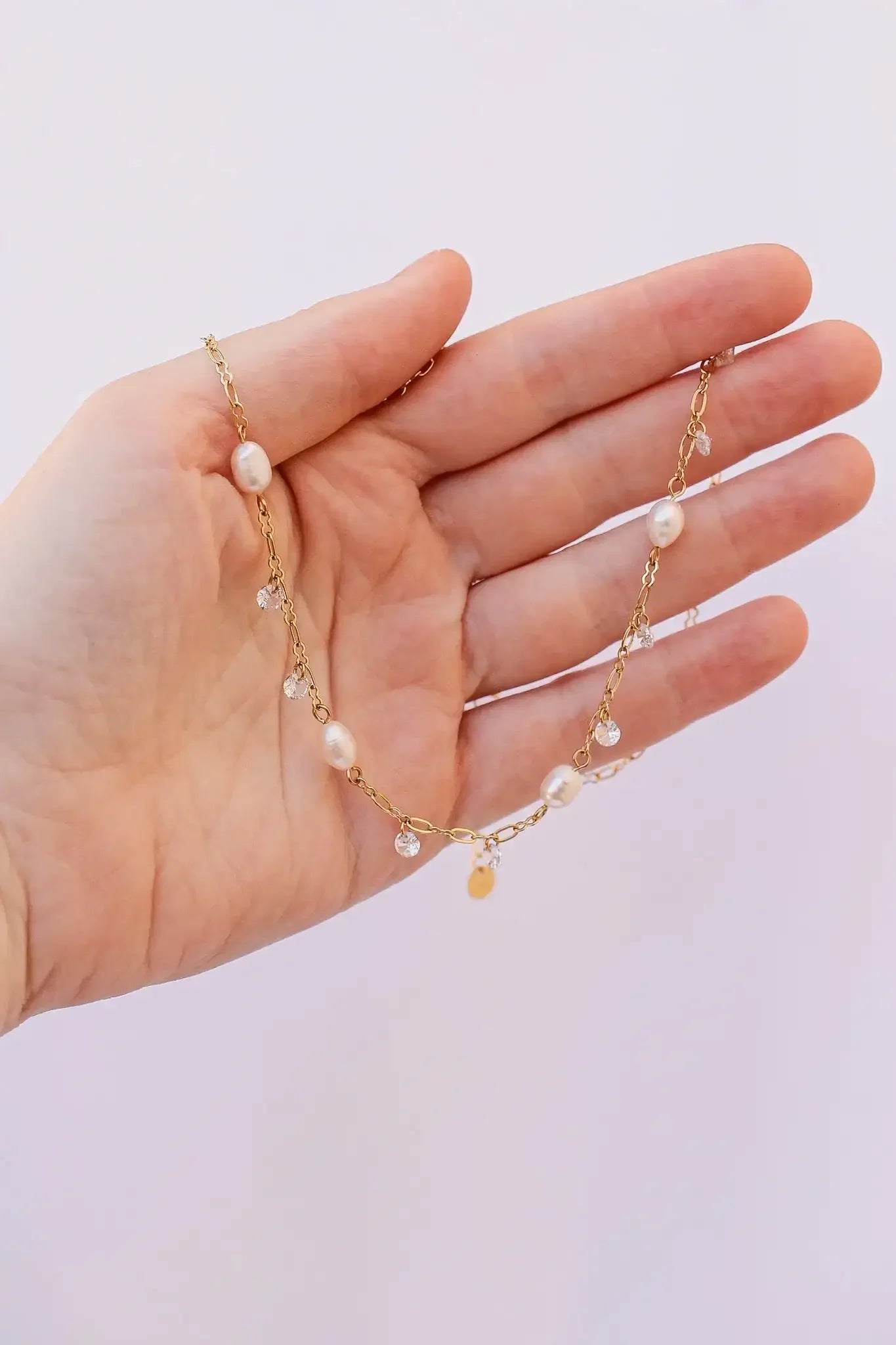 A hand holds a delicate gold necklace adorned with pearls and small clear crystals. The background is a soft, light color, emphasizing the jewelry's elegance.