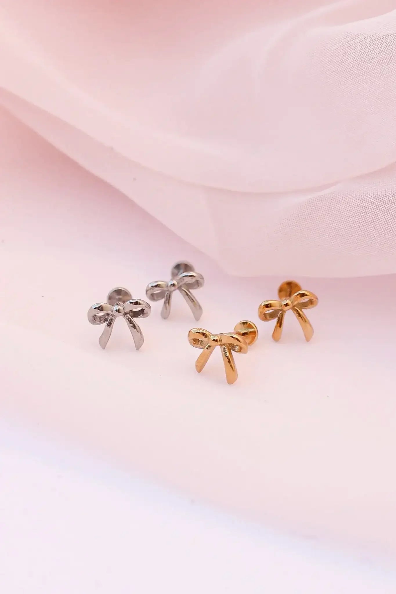 Two pairs of small bow-shaped earrings, one silver and one gold, are displayed on a soft, light pink fabric background, emphasizing their shiny finish and delicate design.
