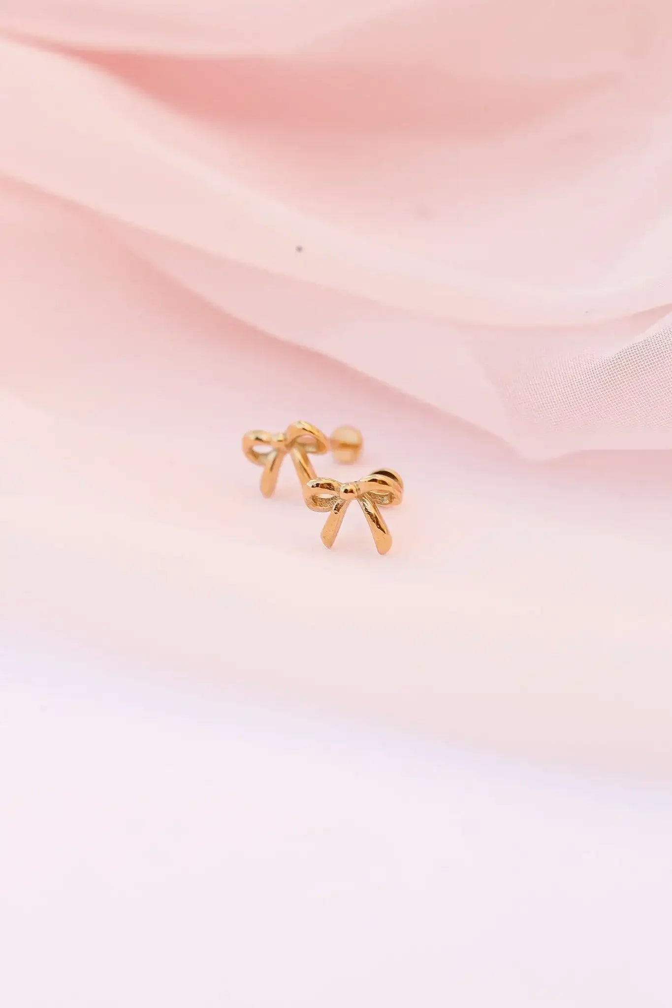 Two small, gold earrings shaped like bows rest on a soft, pale pink fabric. The scene is delicate and feminine, highlighting the jewelry against the textured background.