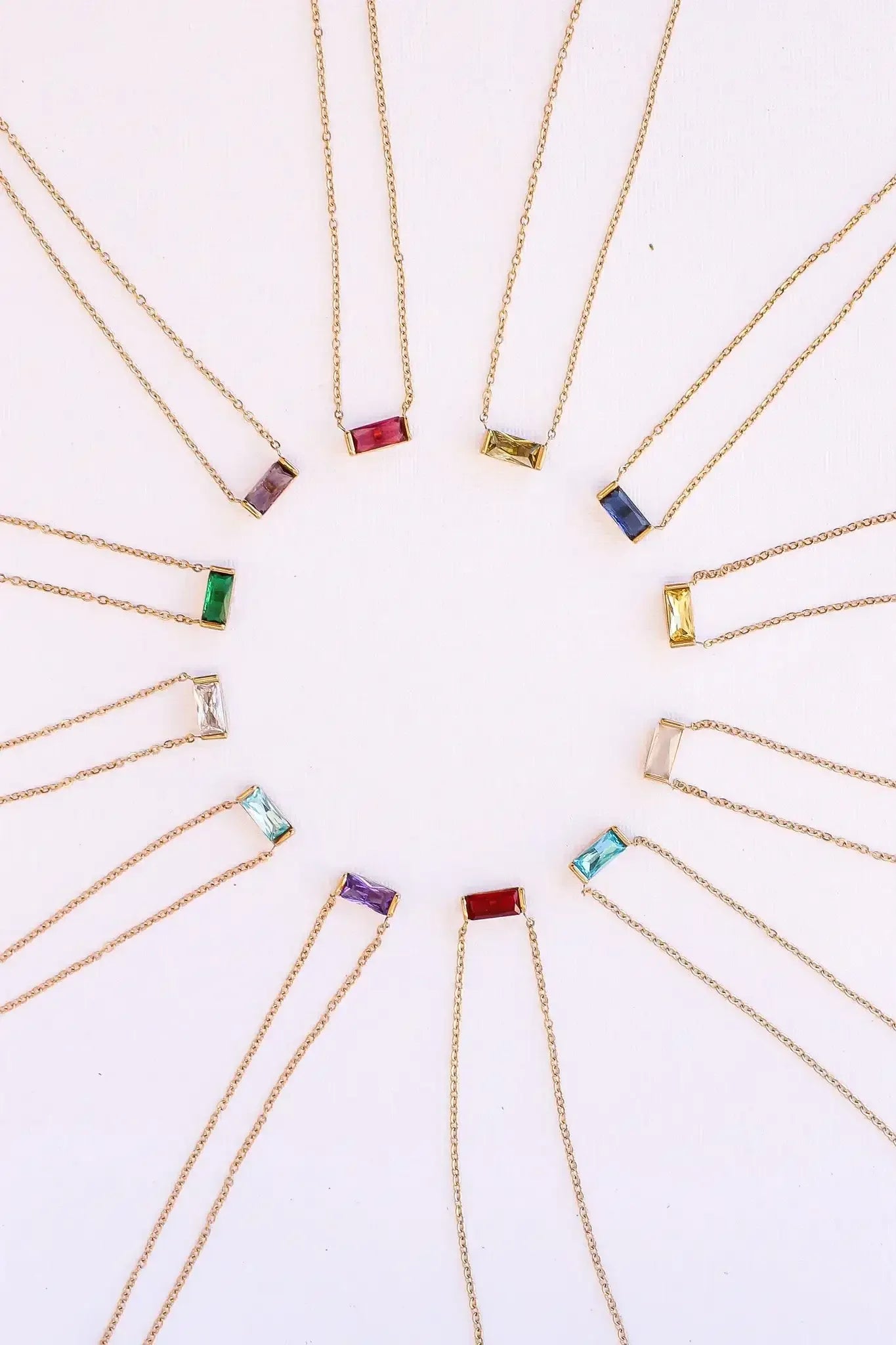 Colorful rectangular gemstone necklaces are arranged in a circular pattern. Each necklace features a gold chain and a different colored stone, displayed on a light-colored background.