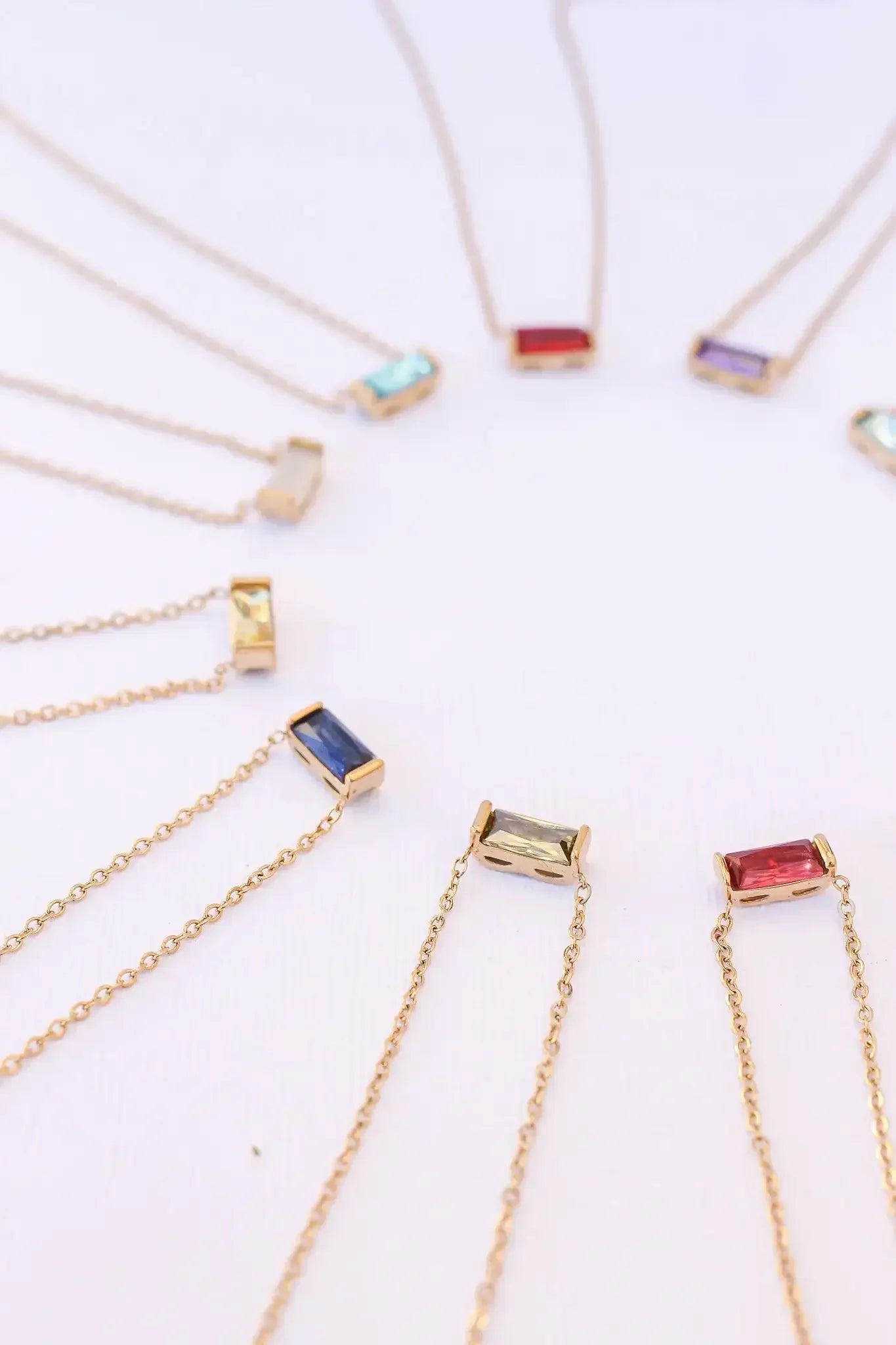 A circular arrangement of delicate gold necklaces with rectangular gemstone pendants in various colors, highlighting their elegance against a light background.