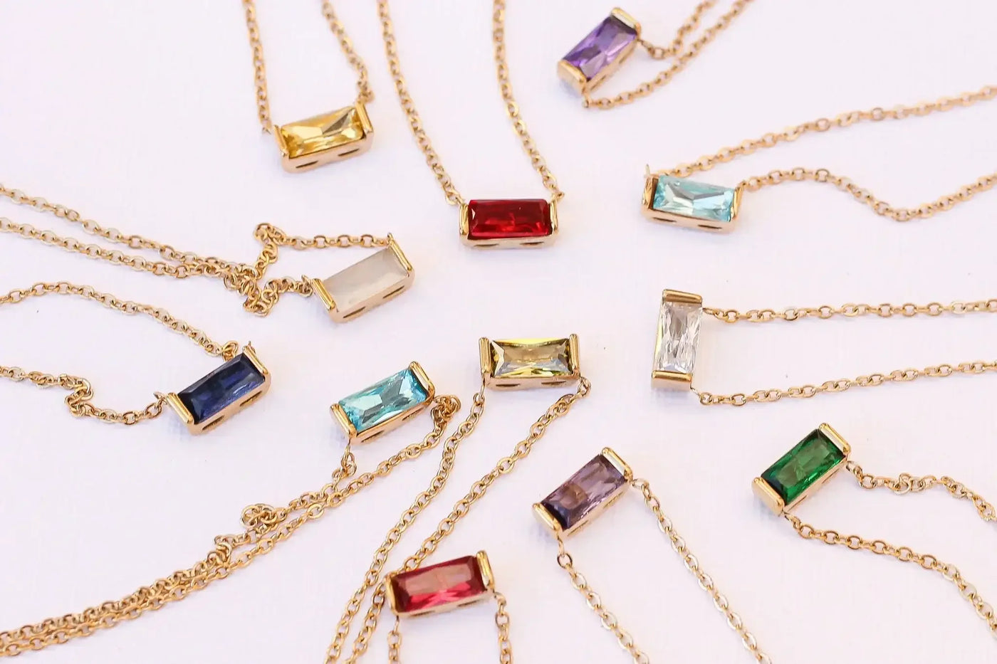 Gold chains with various colored rectangular stones are arranged in a circular pattern on a light background. The stones include shades of blue, green, red, purple, and clear, showcasing their different hues.