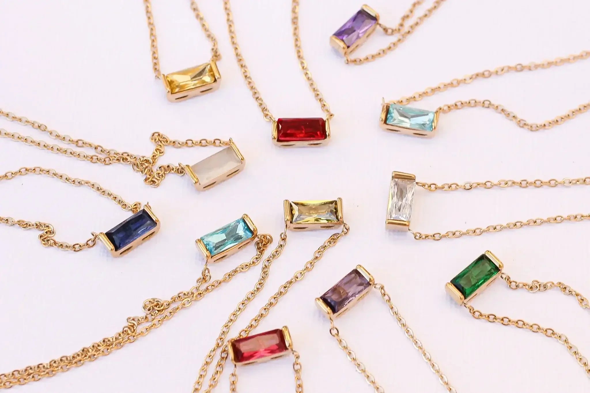 Gold chains with various colored rectangular stones are arranged in a circular pattern on a light background. The stones include shades of blue, green, red, purple, and clear, showcasing their different hues.