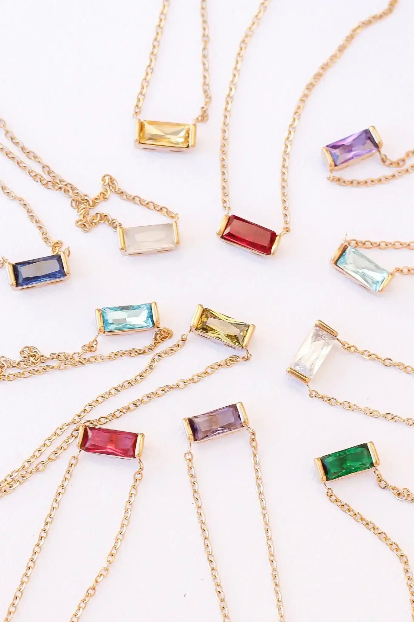A collection of necklaces features various rectangular gemstones in gold settings, displaying colors like red, blue, green, and purple, arranged on a white background.