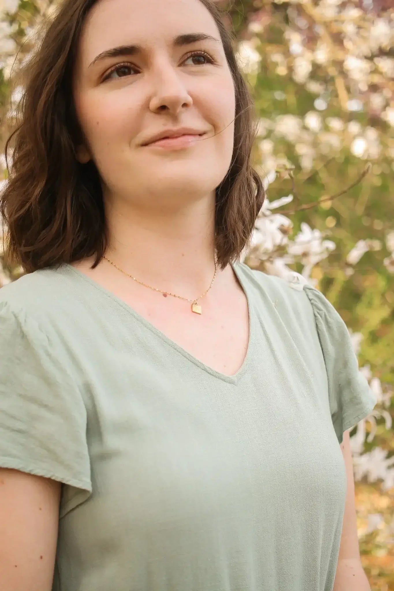 A woman with short, wavy hair looks thoughtfully into the distance. She wears a light green shirt and a delicate gold necklace, surrounded by blooming flowers in a lush garden.