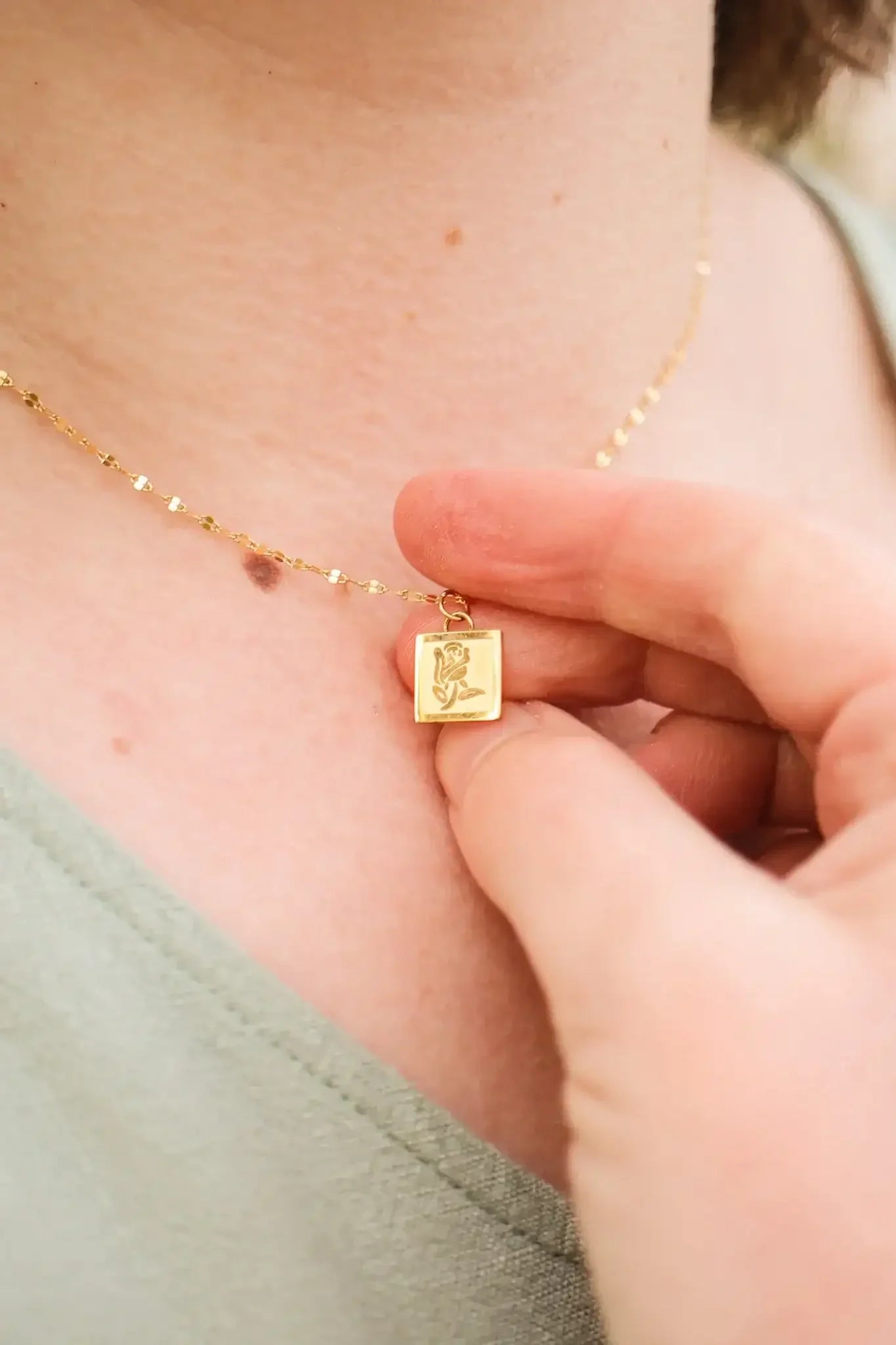 A hand is delicately holding a gold necklace with a small, square pendant featuring a floral design. The pendant rests against a person's skin, with a soft fabric background.
