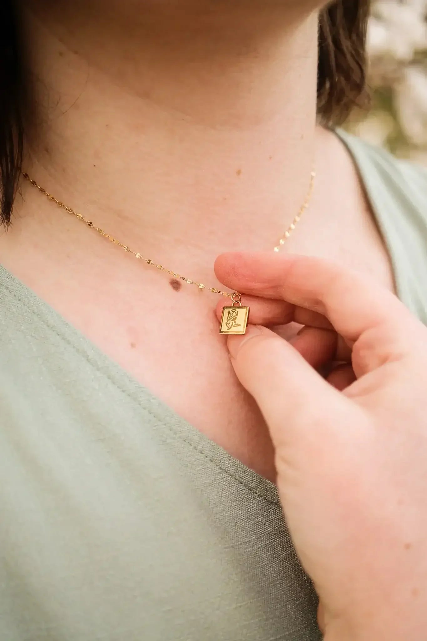 A person is gently holding a gold necklace pendant featuring a floral design. The pendant hangs on a delicate chain, and the individual is located outdoors, surrounded by soft foliage.