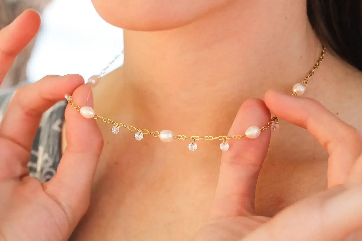A delicate gold necklace adorned with pearls and crystal accents is being showcased by hands near a person's neck, set against a softly blurred light background.