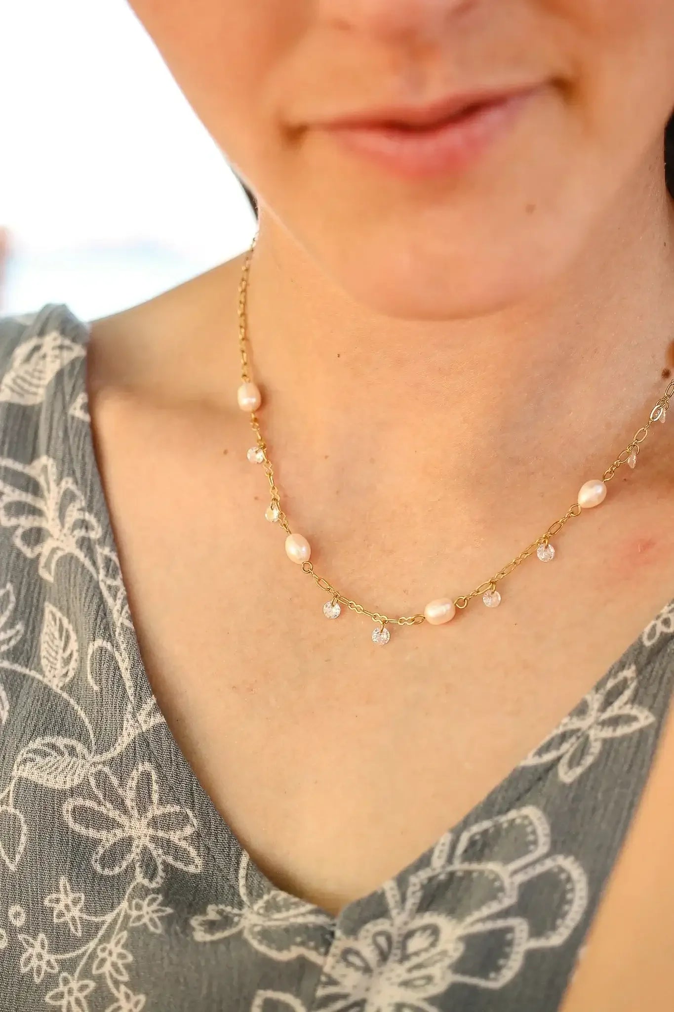 A delicate gold necklace adorned with pearls and crystal drops rests on a woman's collarbone, complementing her floral-patterned garment in a softly blurred background.