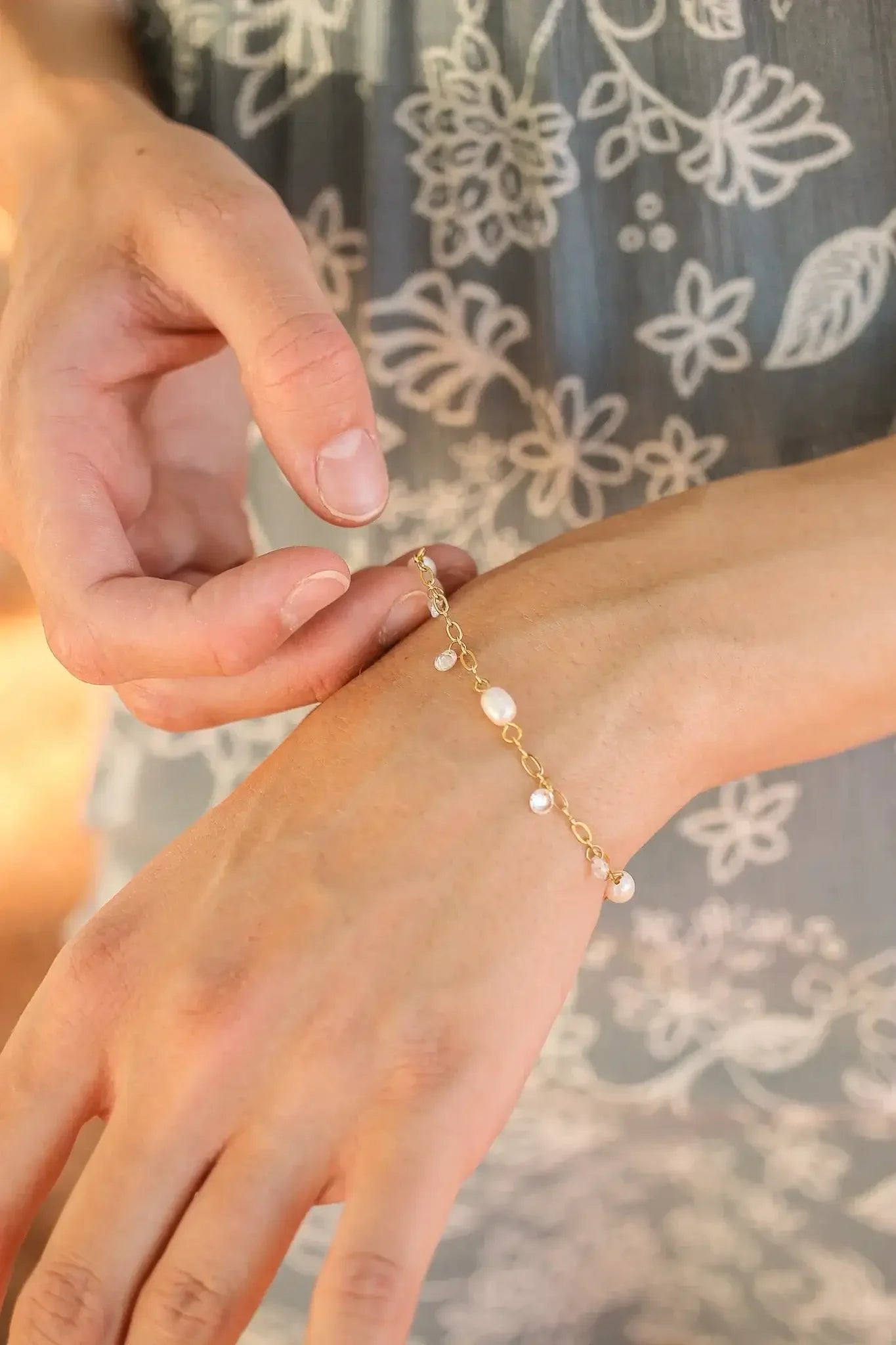 A hand adjusts a delicate gold bracelet adorned with pearls on the wrist. The background features a floral-patterned fabric, suggesting a casual, natural setting.