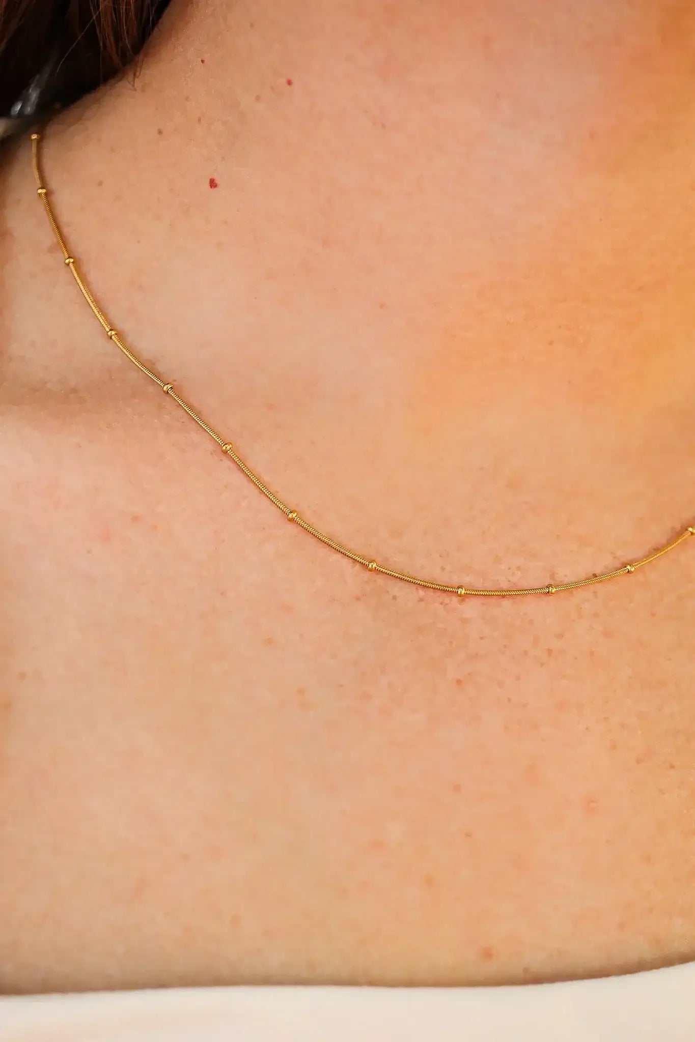 A delicate gold necklace rests on bare skin, with small beads evenly spaced along the thin chain, showcasing a subtle shine against a smooth, lightly tanned surface.