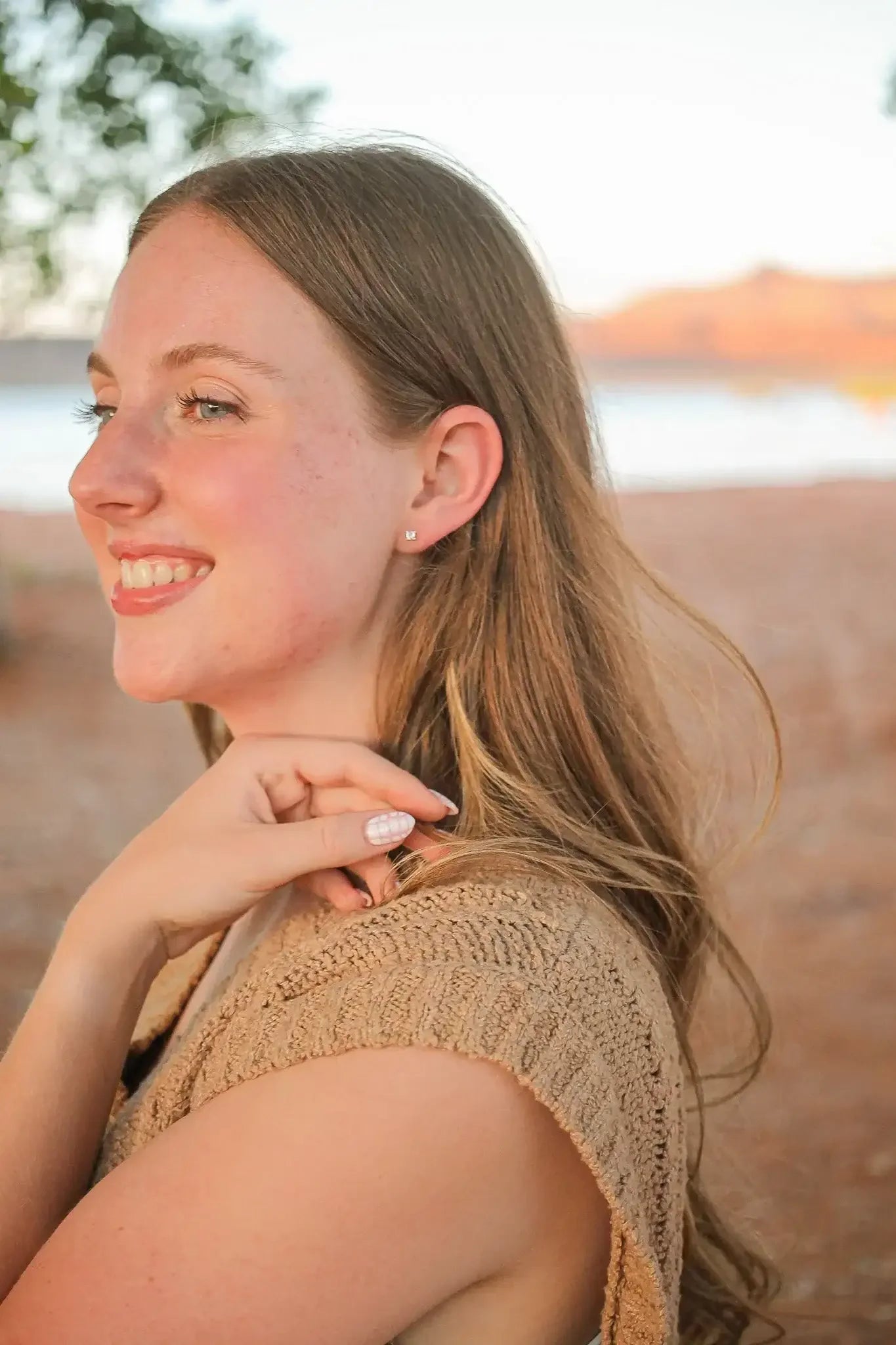 A young woman smiles while touching her hair, wearing a sleeveless, textured beige top. She stands in a natural setting, with a blurred water body and hills in the background.