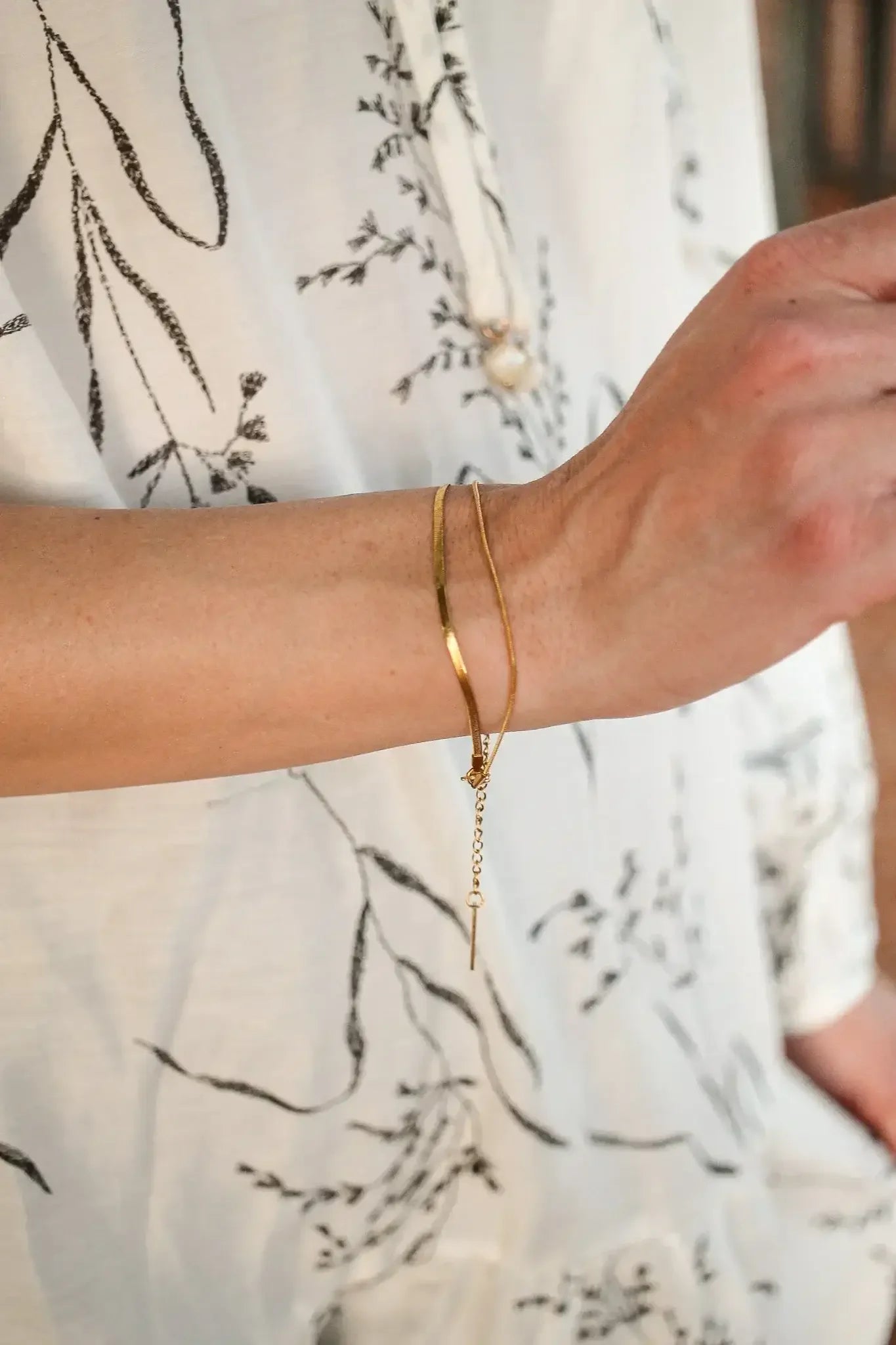 A hand wearing two gold bangles is raised, showcasing the jewelry's reflective surface against a light, patterned blouse. The environment is softly illuminated, enhancing the elegant appearance.