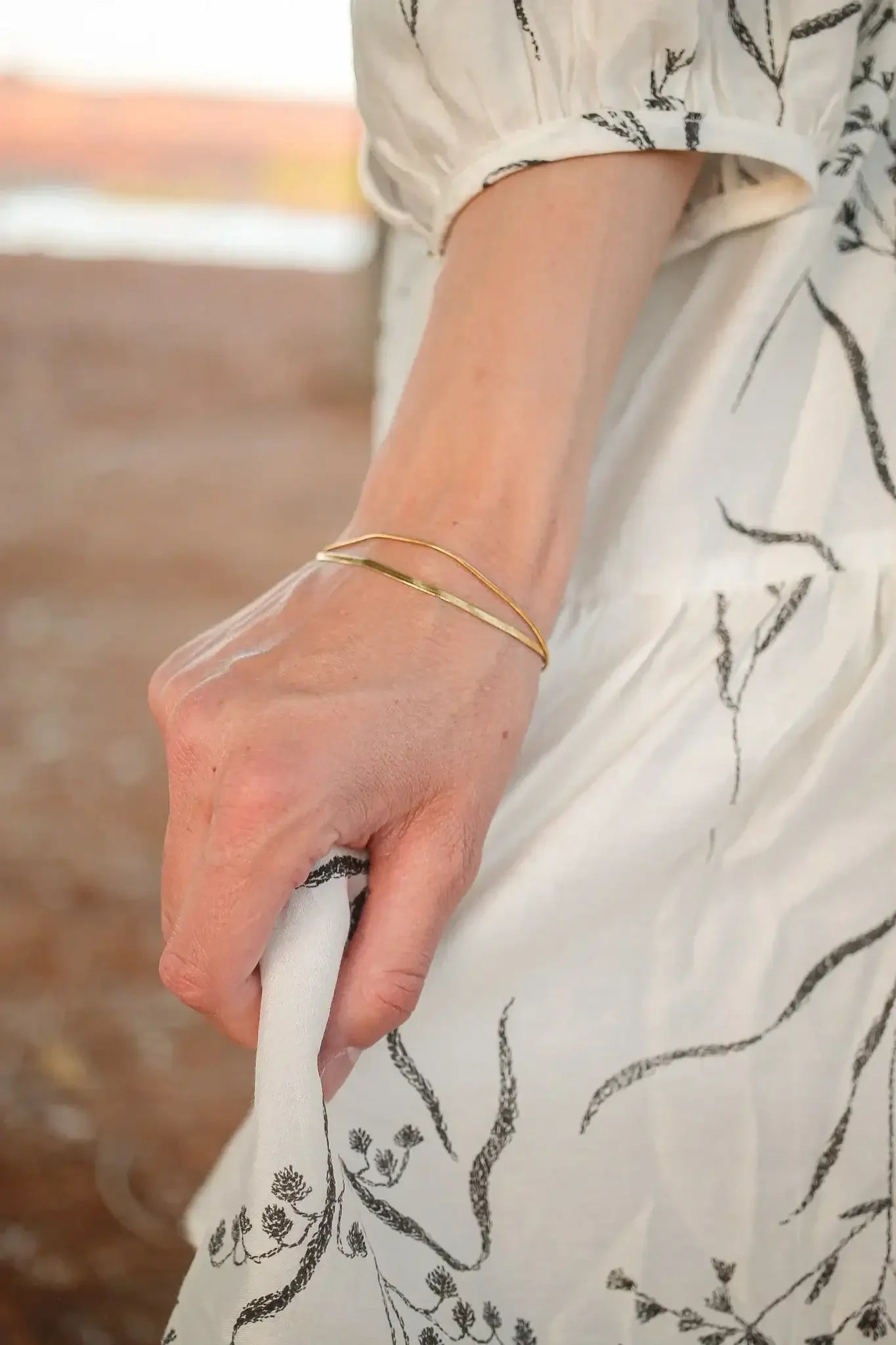 A hand grips the hem of a white dress with a black floral pattern, showcasing a thin gold bracelet on the wrist. The setting appears natural and rustic.