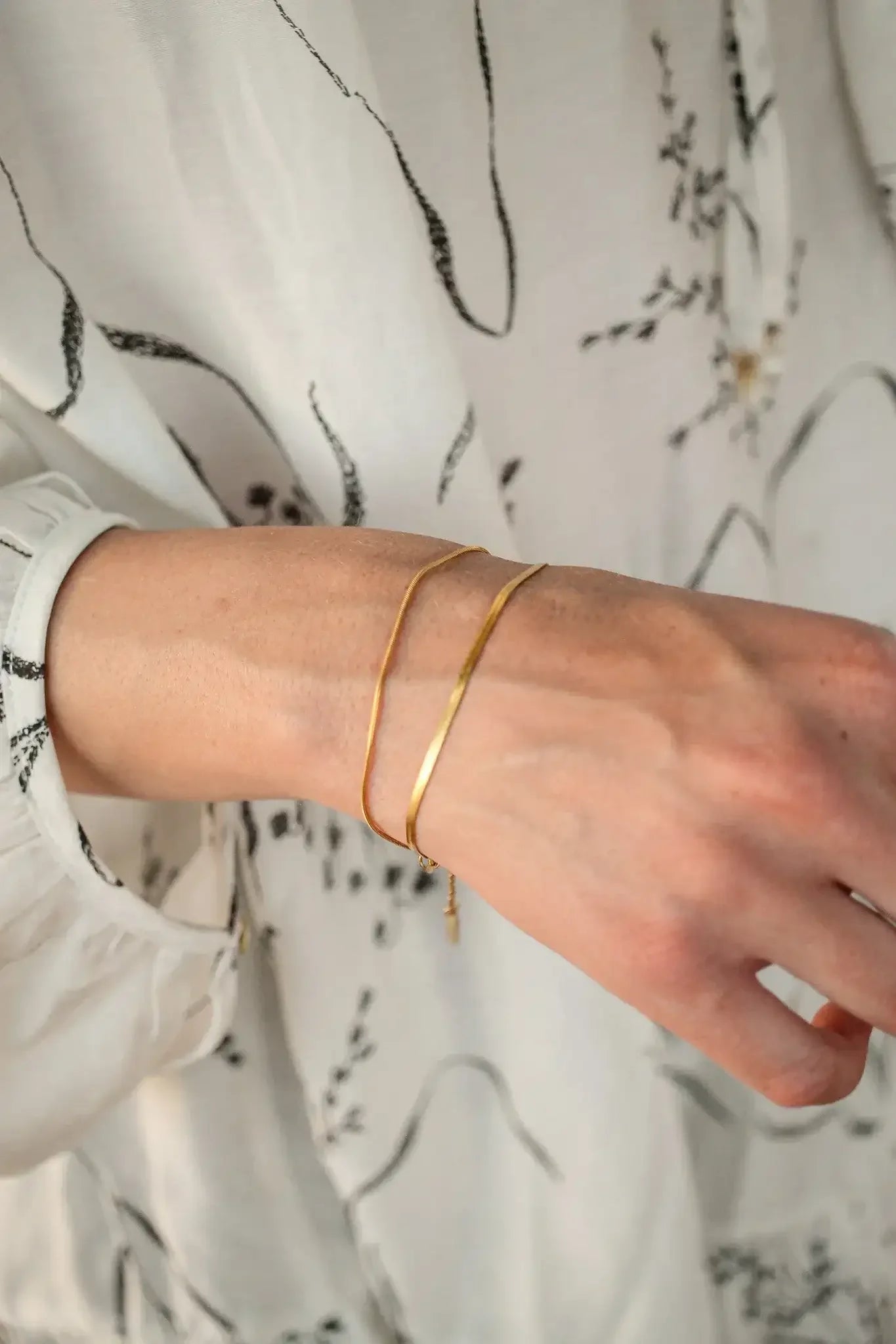 A hand adorned with two thin gold bracelets extends from a white blouse with black floral patterns, highlighting the jewelry against a soft, light background.