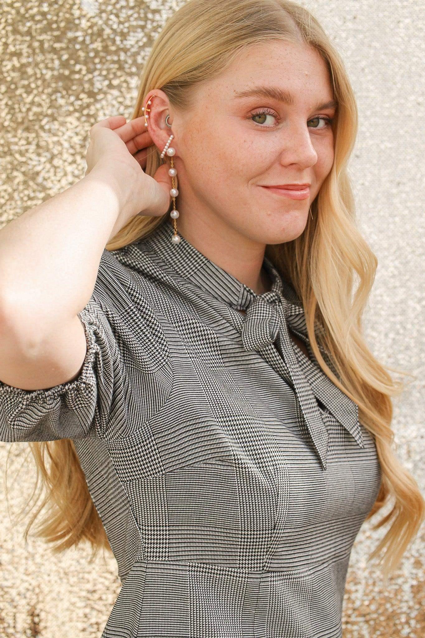 A person with long blonde hair smiles while holding an earring, showcasing it. They wear a patterned shirt, with a glittering golden background creating a festive atmosphere.