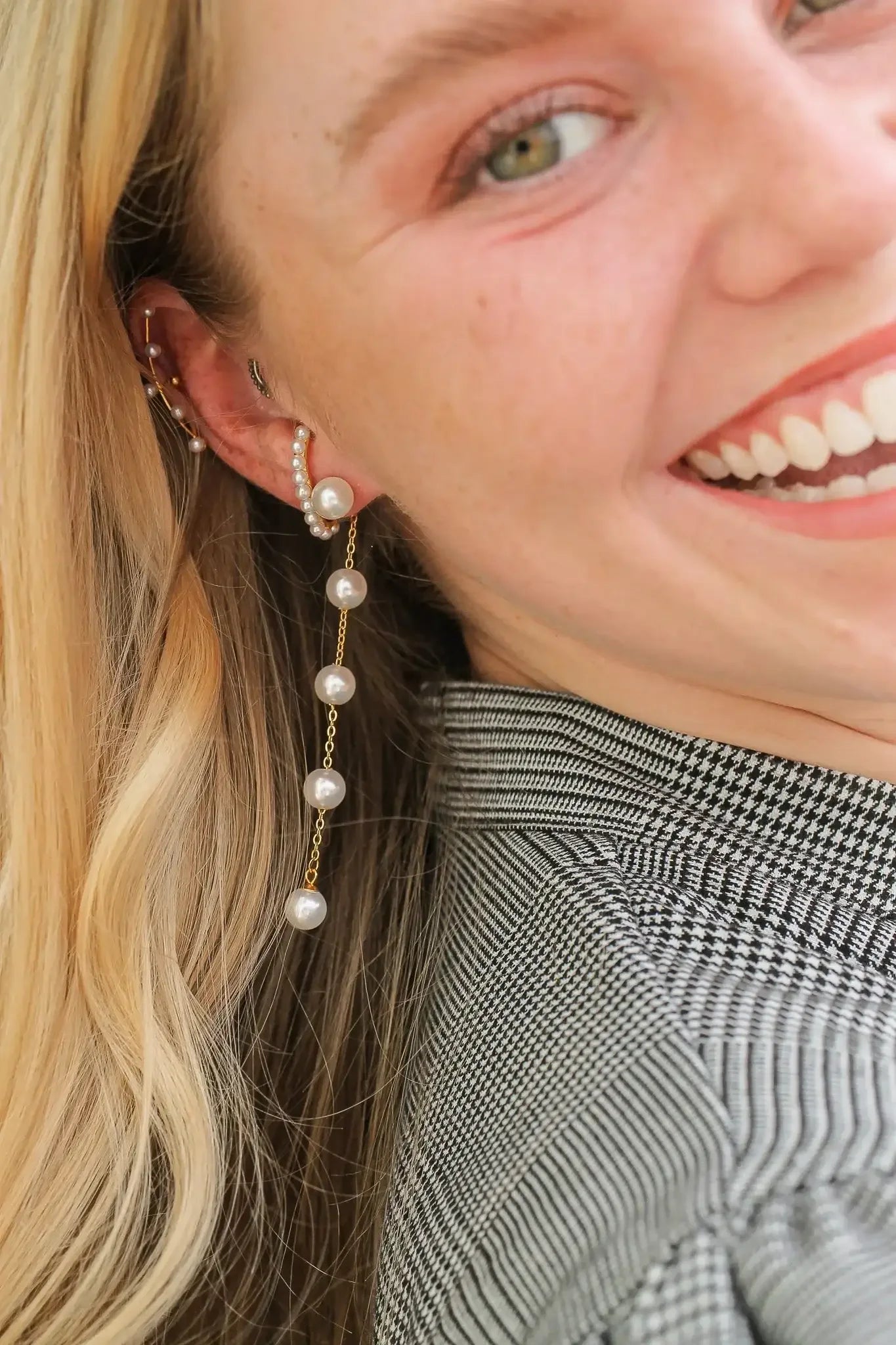 A woman smiles, showcasing a cascading pearl earring. The earring, attached to her ear, features pearls linked by delicate gold chains. She wears a patterned shirt, with her hair falling loosely.