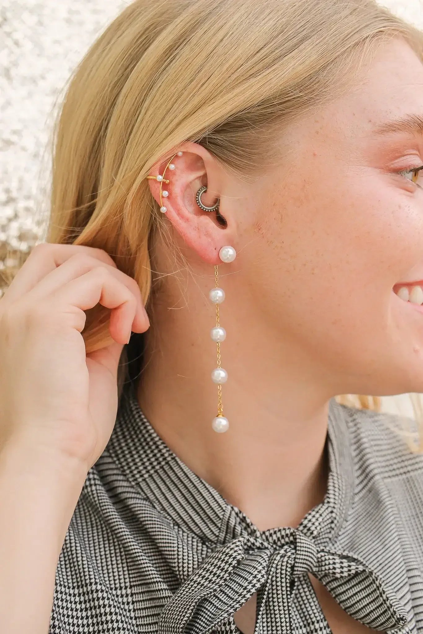 A woman with blonde hair is smiling while touching her ear. She wears a long pearl earring and multiple ear piercings, against a softly blurred, light background.