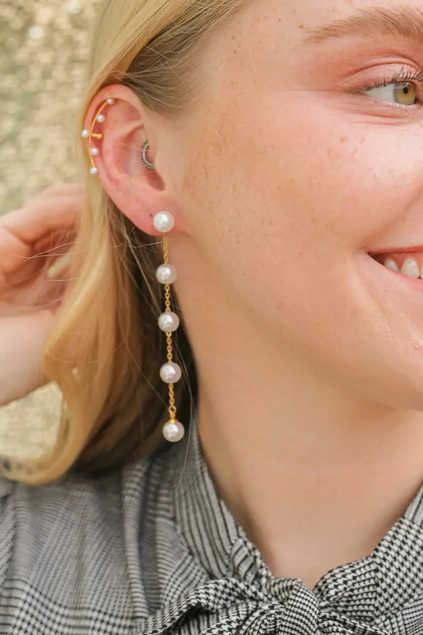 A woman with long blonde hair smiles while holding her hair back. She wears a long pearl earring that dangles down, complementing her plaid shirt and glowing skin.