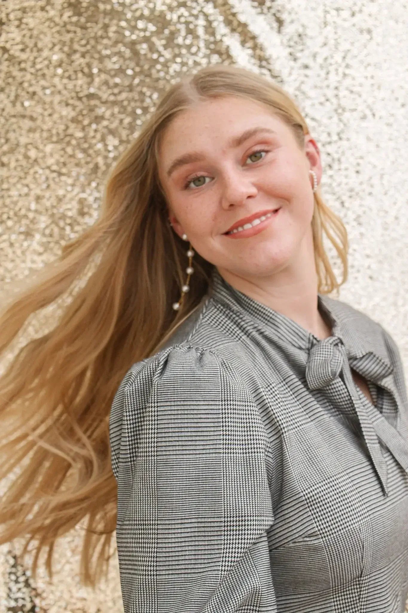 A young woman with long, flowing blonde hair smiles as she turns her head, wearing a black and white checkered blouse against a shimmering gold backdrop.