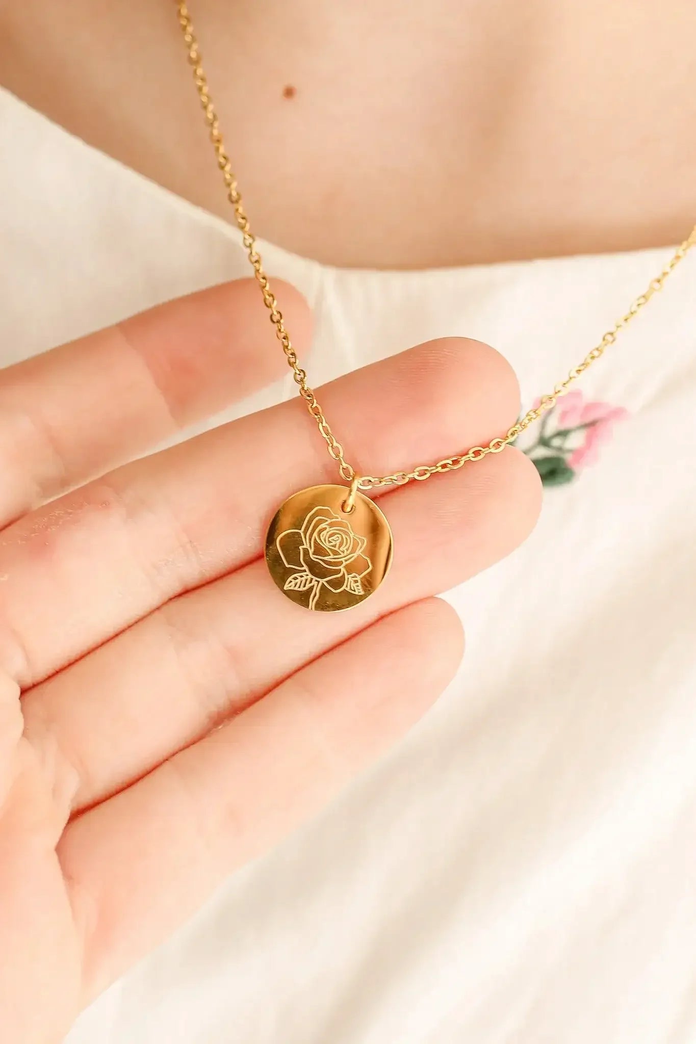 A hand is holding a gold necklace featuring a circular pendant with an engraved rose design. The background shows a light-colored fabric with a small pink flower embroidery.