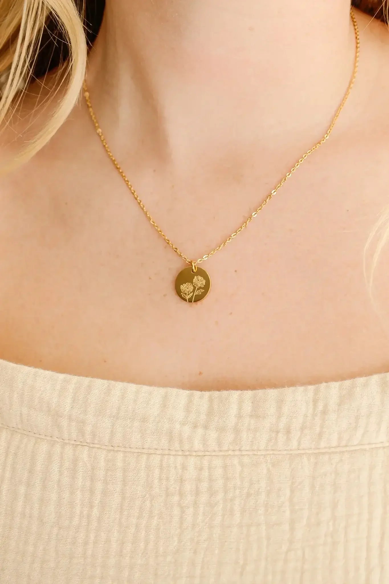 A gold necklace with a circular pendant featuring an engraved floral design rests on a personâ€™s neck, which is partially covered by a textured, light-colored top.