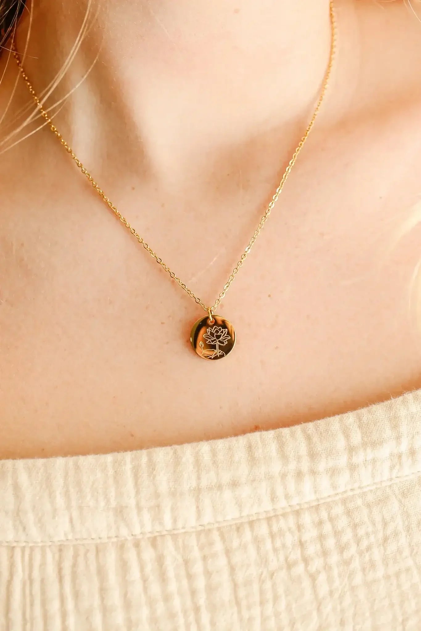 A gold necklace featuring a circular pendant with a floral engraving rests on a person's collarbone, surrounded by soft fabric in a neutral tone.