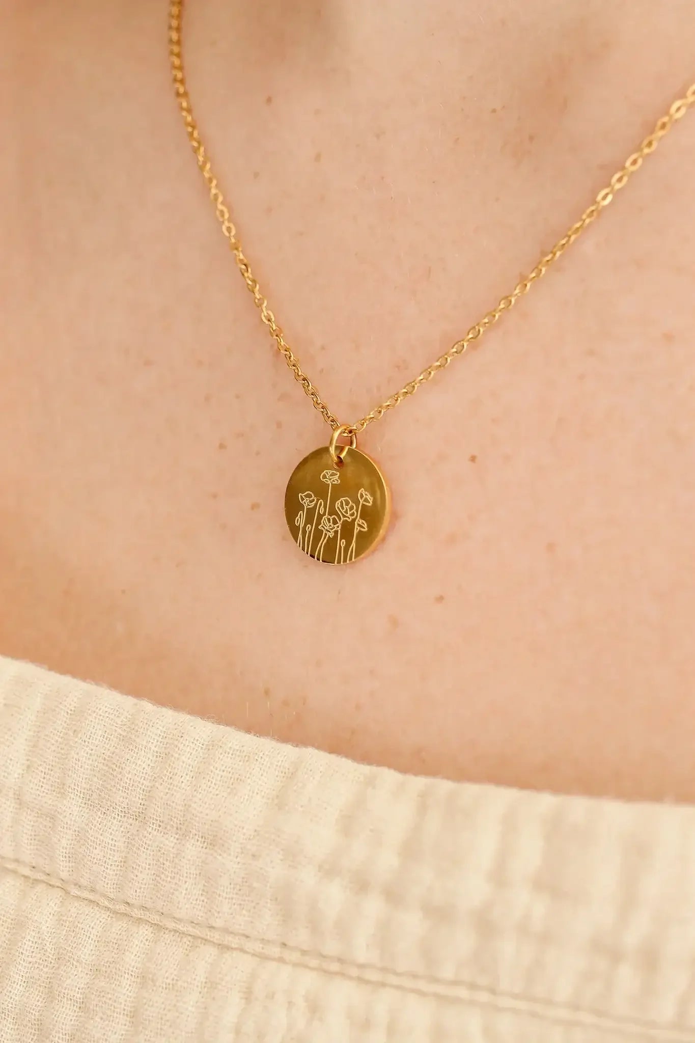 A gold chain necklace features a round pendant engraved with floral designs. It rests on a person's bare neck, which is partially covered by a light, textured fabric.