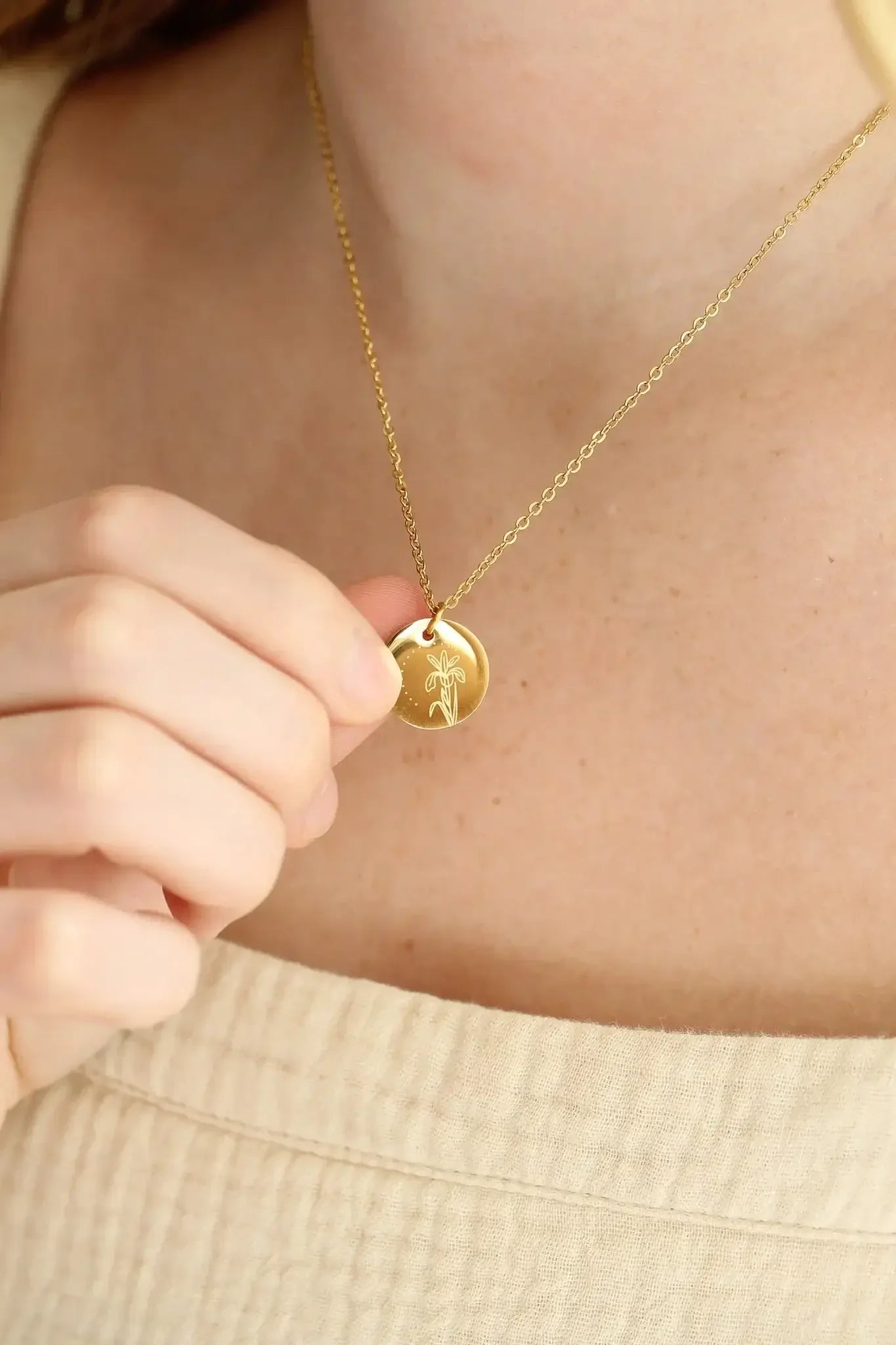 A person holds a gold pendant necklace featuring a floral design, displaying it against a softly lit, neutral-toned background that reveals part of their skin and clothing.