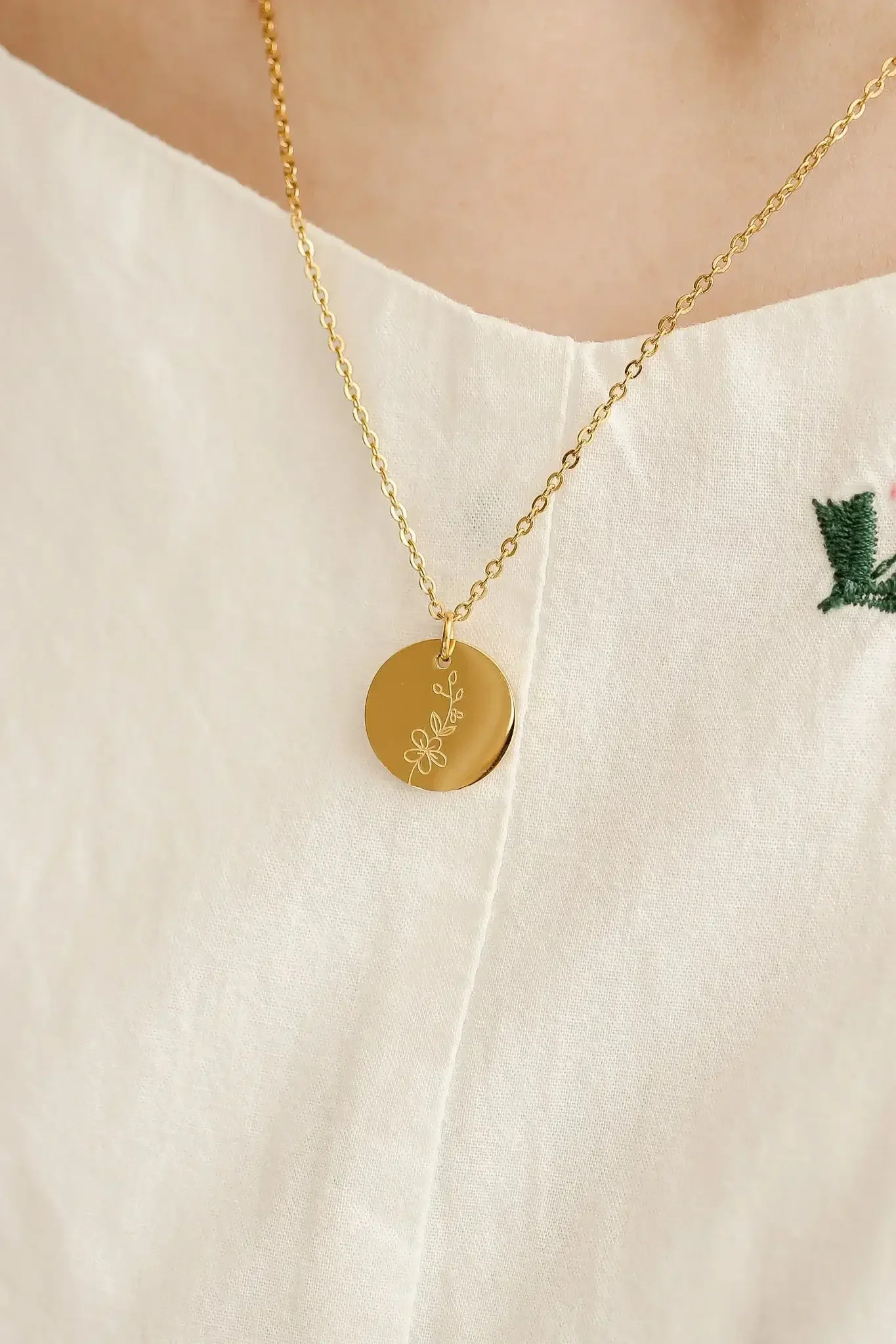 A gold necklace with a circular pendant featuring a floral design hangs on a person's neck, showcasing delicate chain links against a light-colored, textured garment.