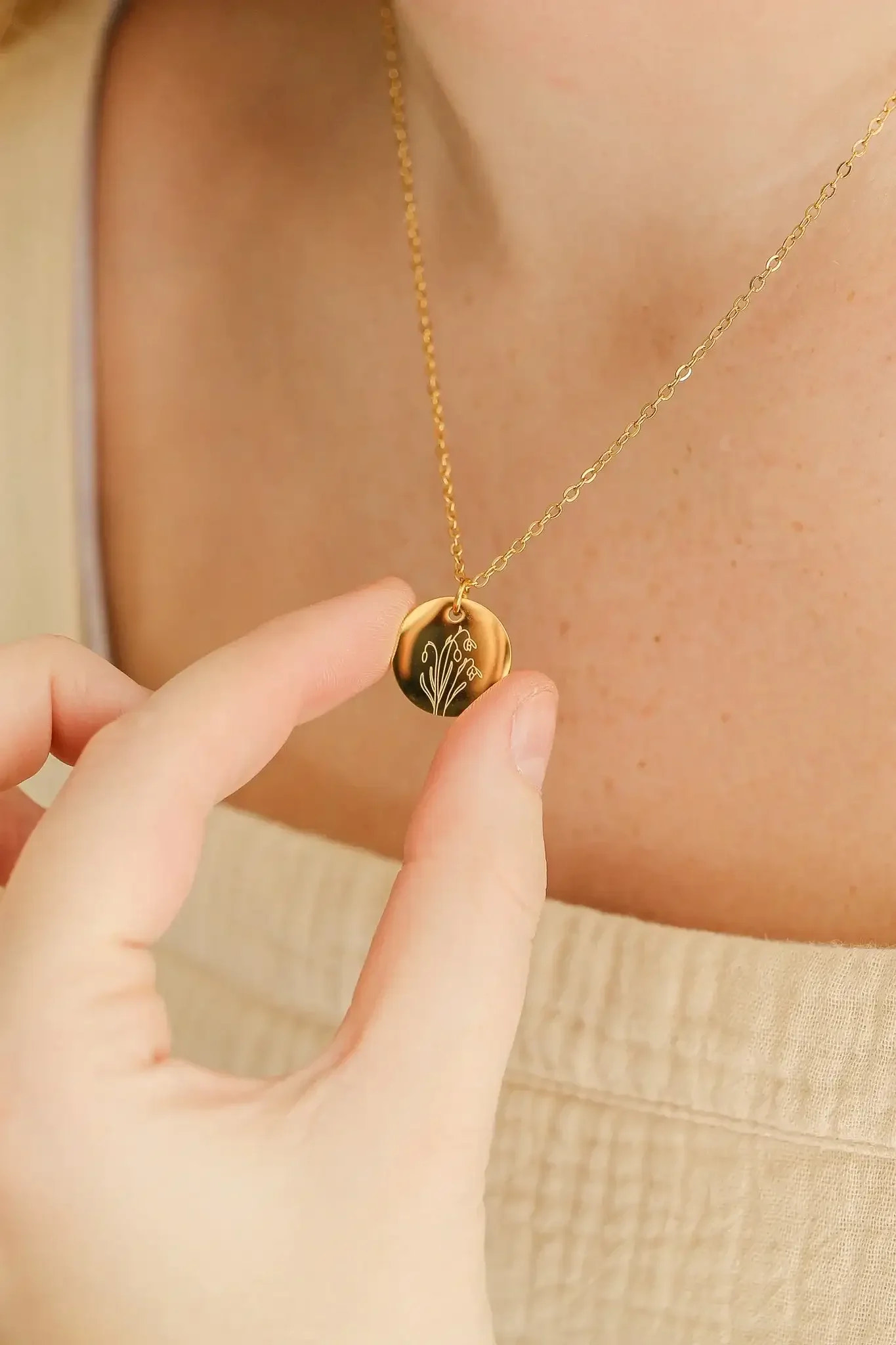 A hand holds a gold pendant necklace, which features a floral design, close to the neck of a person wearing a light-colored, textured top. The skin is lightly freckled.