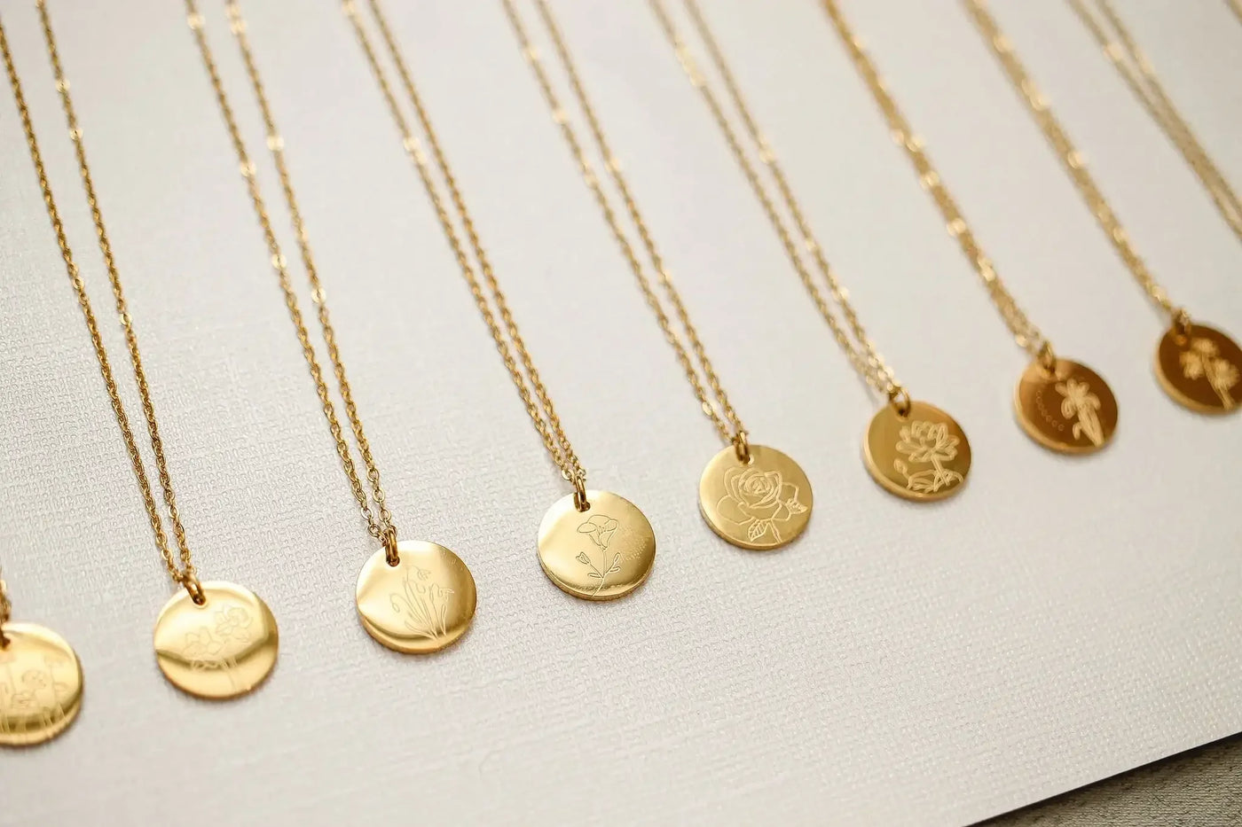 Gold necklaces hang in a row with engraved circular pendants, showcasing various floral designs. They are displayed on a textured light-colored background, emphasizing their elegance.