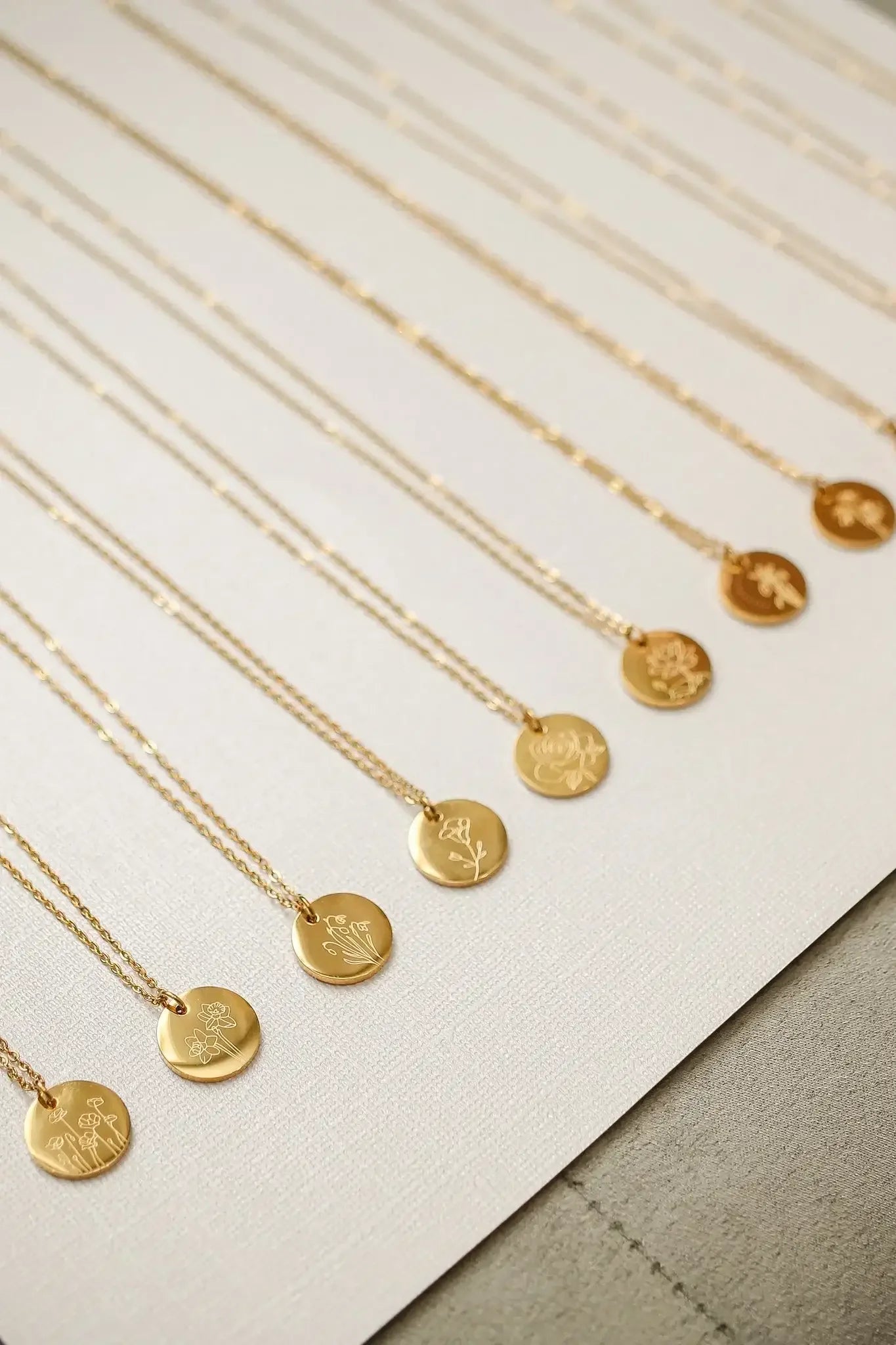 Gold necklaces hang in a straight line on a textured surface. Each necklace features a circular pendant engraved with floral designs, showcasing an elegant display of jewelry.