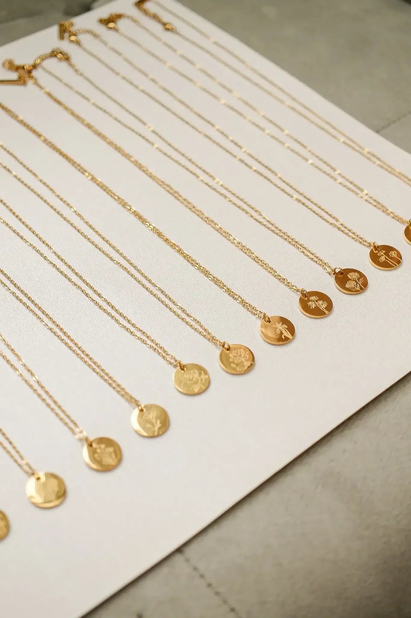 Gold necklaces with circular pendants, some engraved, are neatly arranged on a light fabric background. The jewelry is presented in a way that highlights their intricate designs and varying lengths.