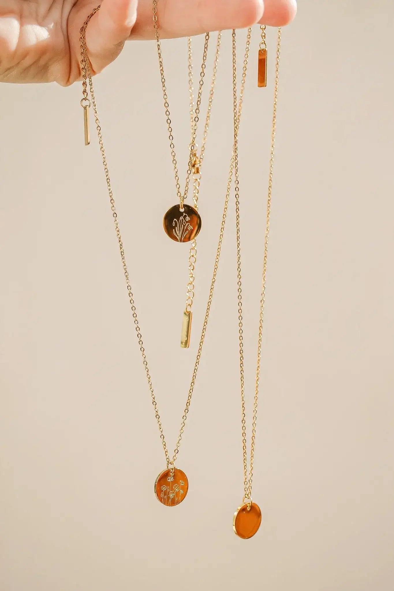 A hand holds several gold necklaces, showcasing various pendants including circular ones with floral designs and small rectangular charms, against a soft, neutral background.