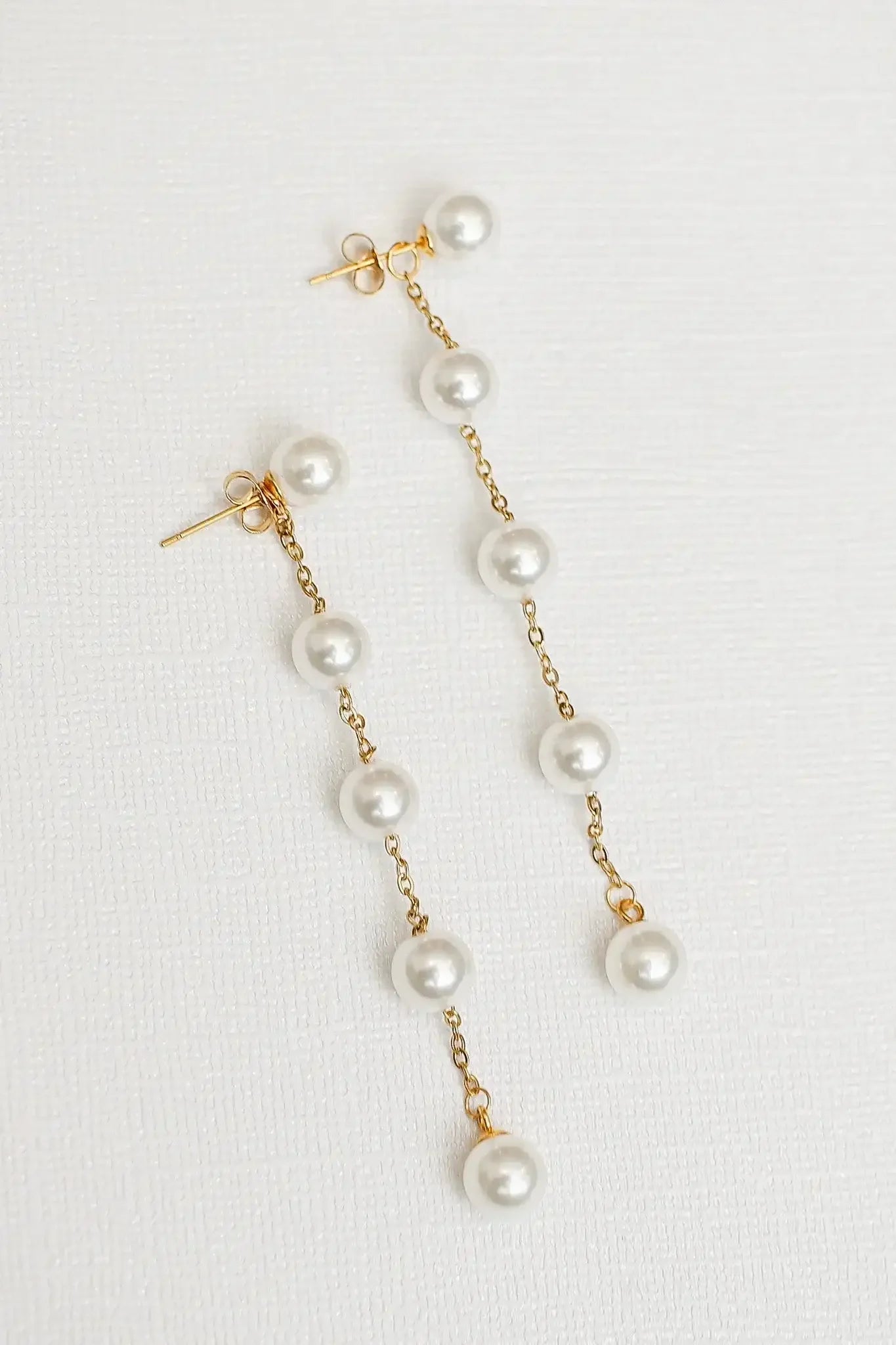 Dangling earrings feature a series of glossy pearls linked by delicate gold chain. They are positioned against a plain, textured white background, emphasizing their elegant design.