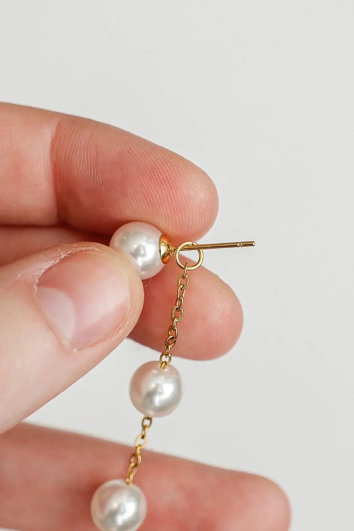 A hand holds a chain earring with multiple pearls, positioned for insertion into an ear. The background is plain and light, emphasizing the jewelry.