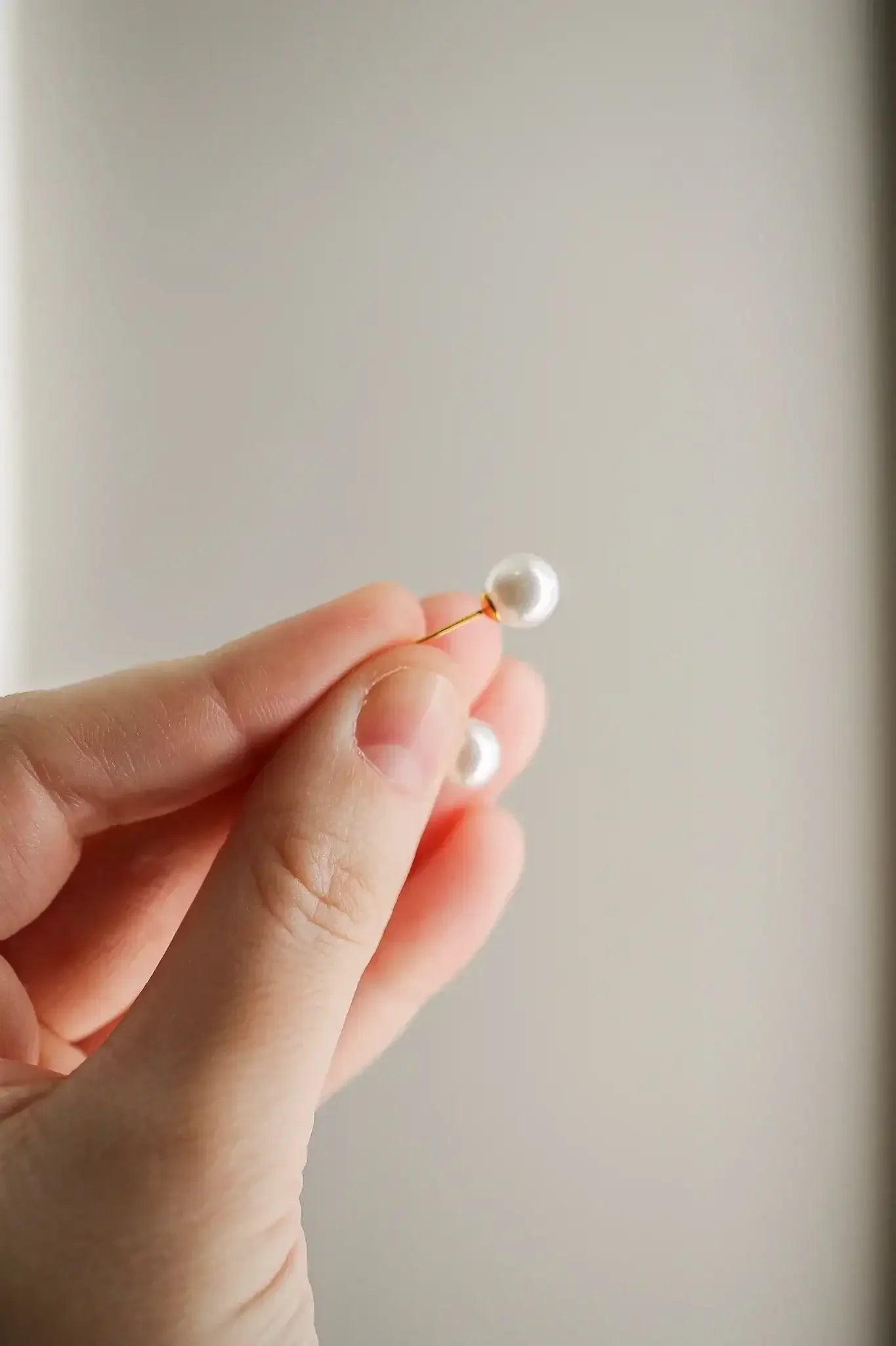 A hand holds a thin wire with a pearl attached. The action suggests preparation for jewelry making. The background is a soft, neutral color that emphasizes the pearl's luster.