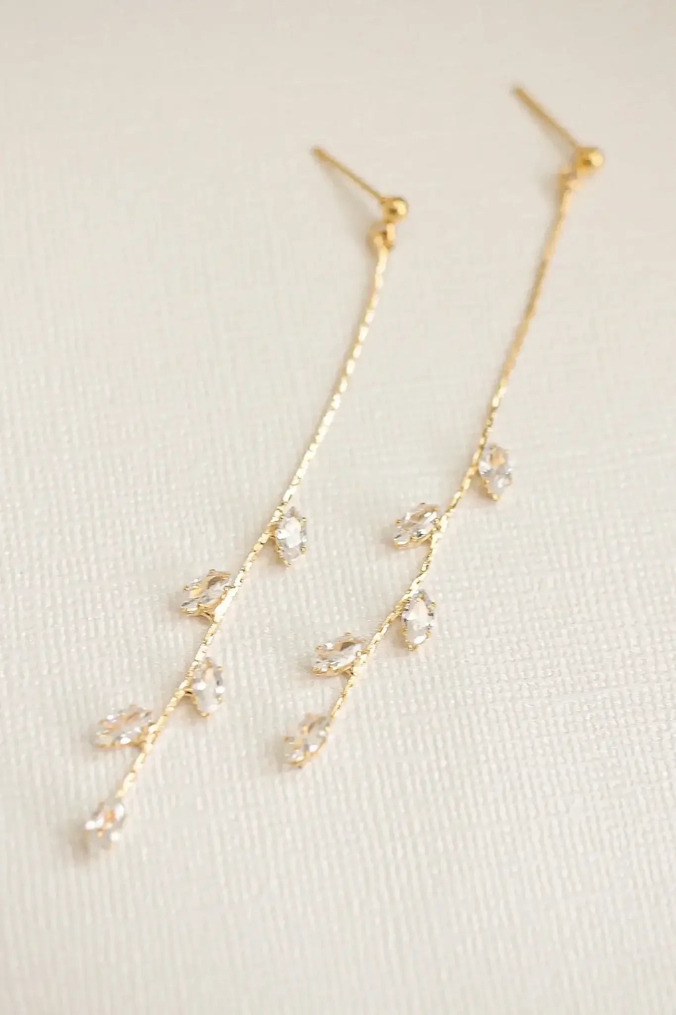 Two gold earrings with delicate, vine-like designs hang gracefully. They feature small, sparkling crystals arranged in a linear pattern. The background is a simple, textured cream surface.