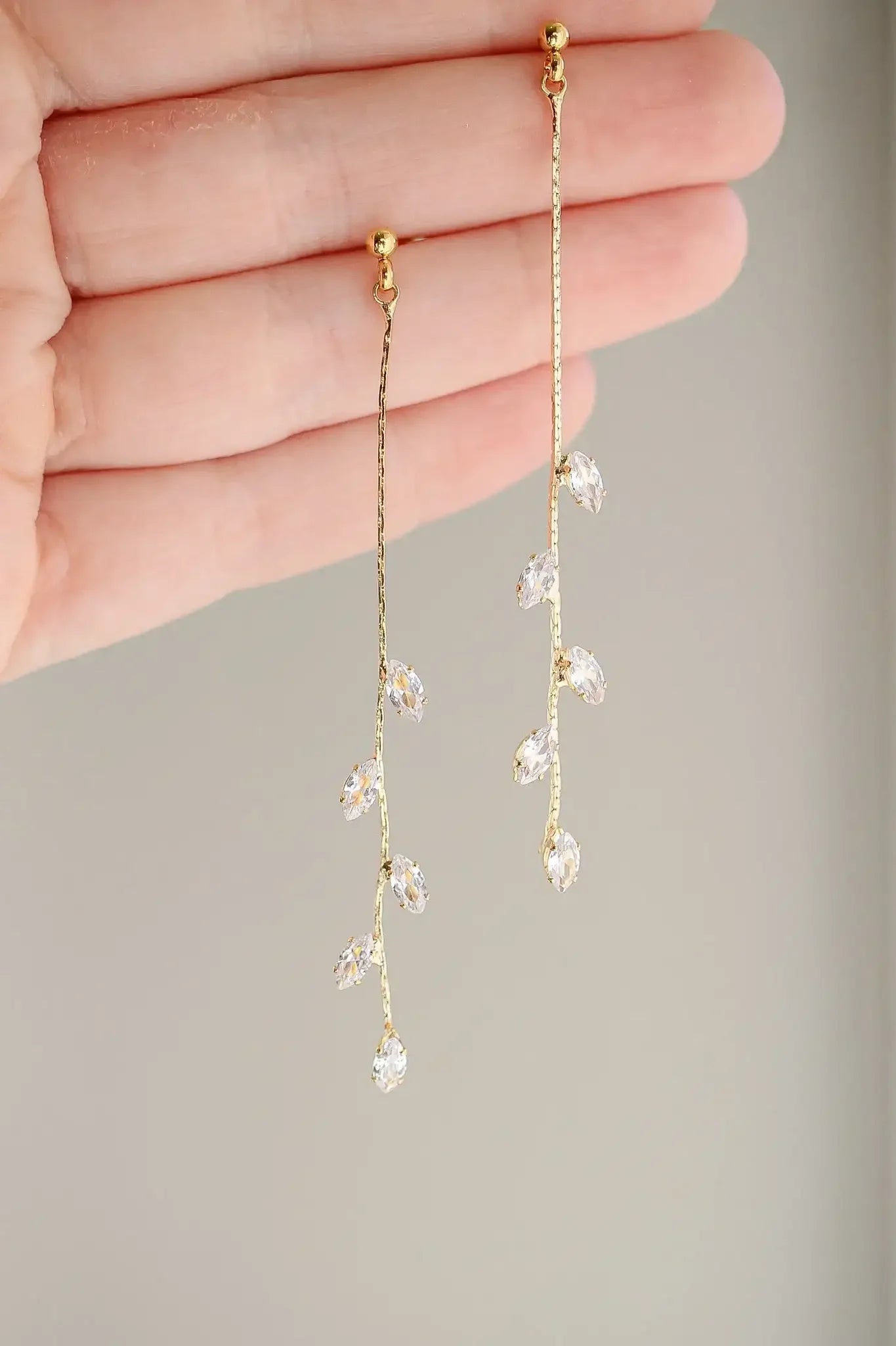 Dangling earrings with a gold chain, featuring clear crystal accents resembling leaves, are being held up by a hand against a soft, neutral background.