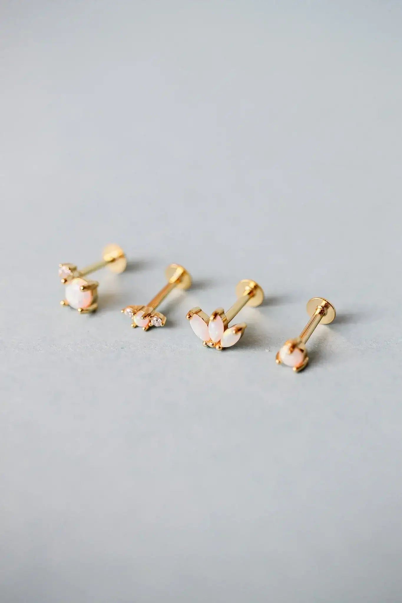 Gold earrings with opal-like stones are arranged in a row on a smooth, light gray surface. Their elegant designs vary, showcasing floral and geometric patterns.