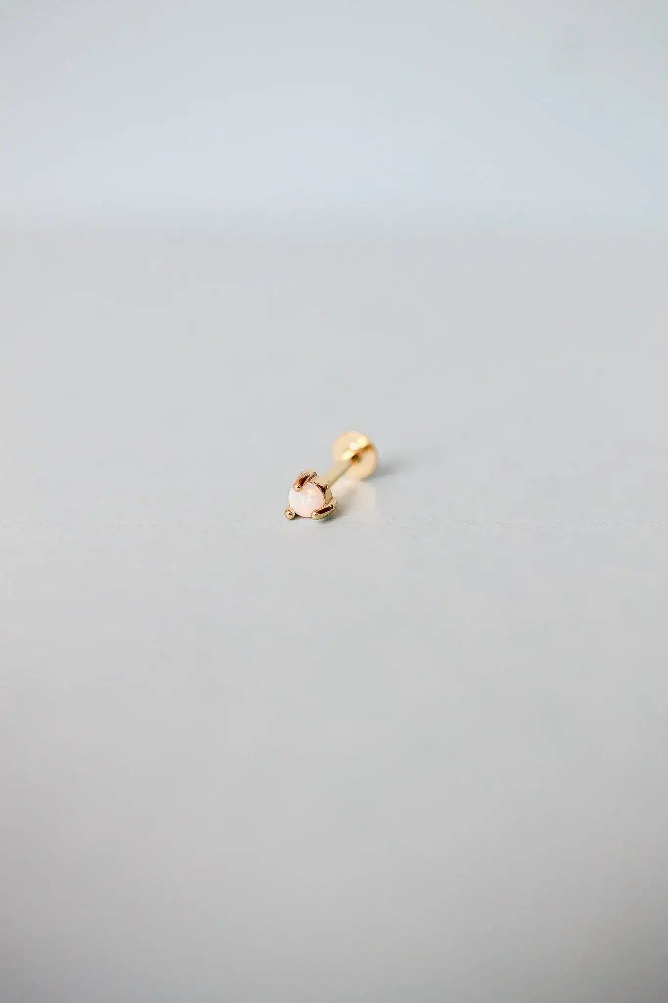 A small gold earring lies on a light gray surface, featuring a round opal stone at the end. The setting has a delicate design, suggesting elegance in its simplicity.