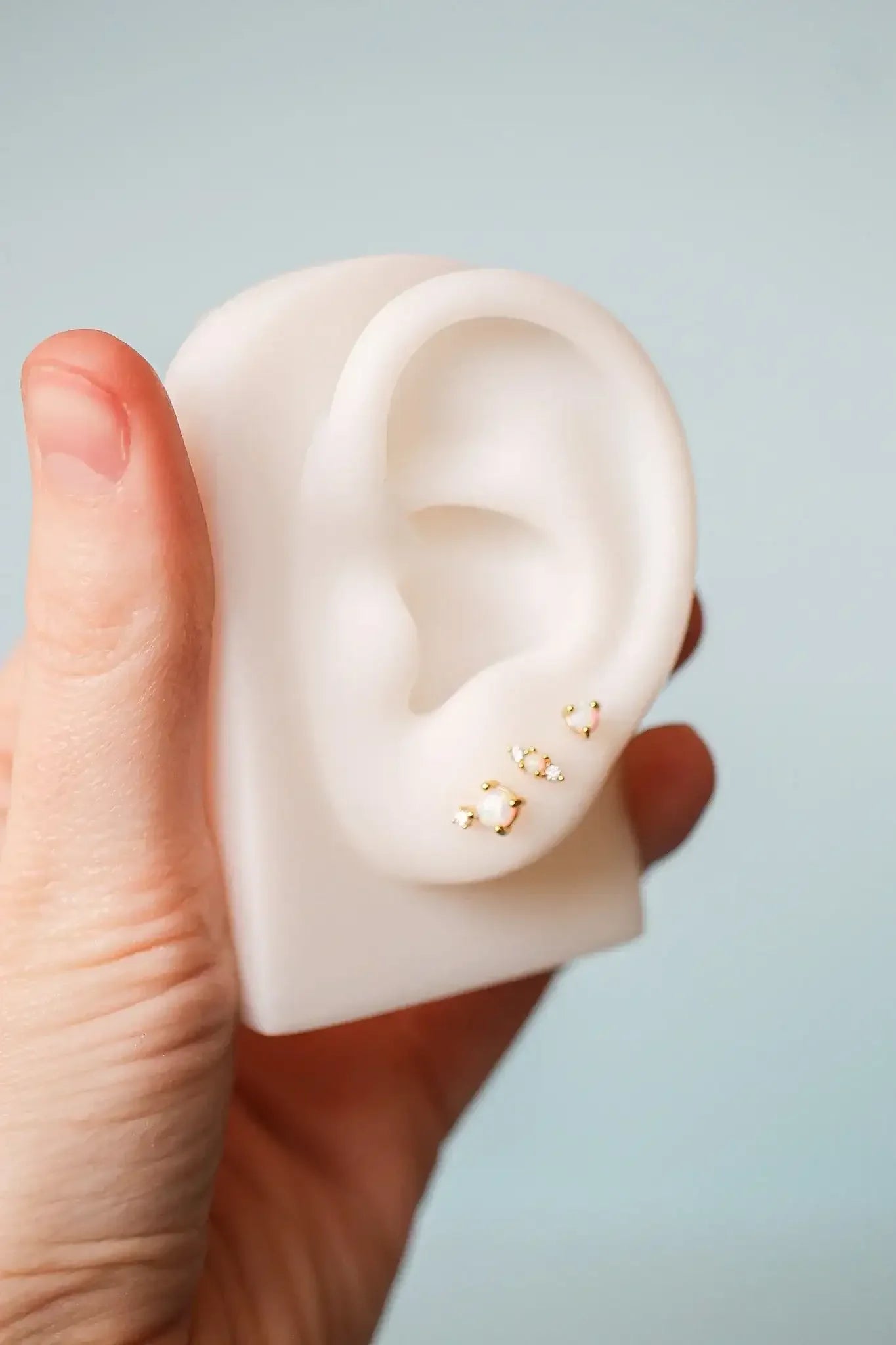 A person holds a realistic, white ear model decorated with several small, sparkling earrings. The background is a soft, light blue, emphasizing the delicate display of accessories.