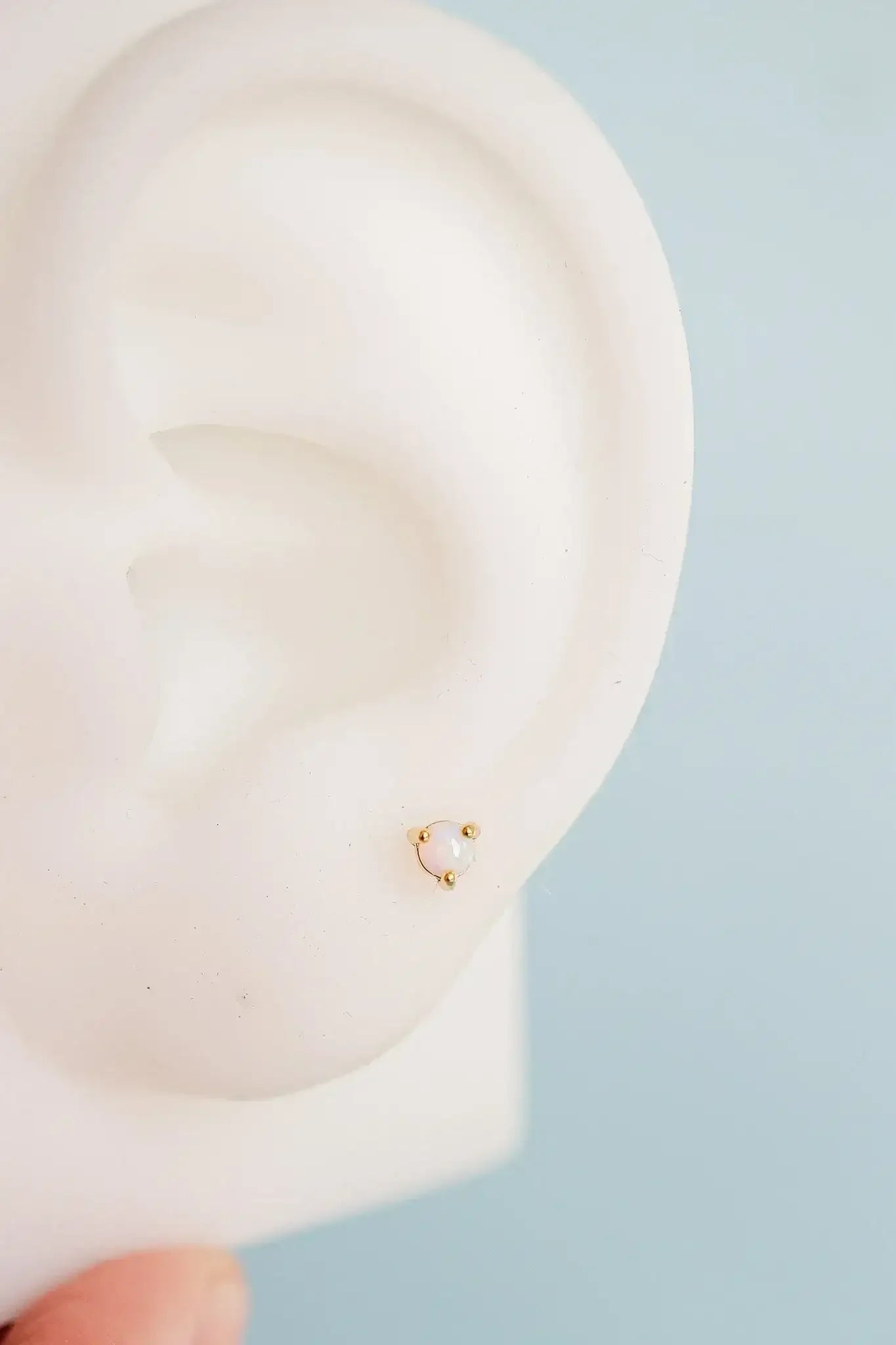 A small, delicate earring featuring a pearl is displayed on a lifelike ear model. The background is a soft blue, enhancing the jewelry's appearance.