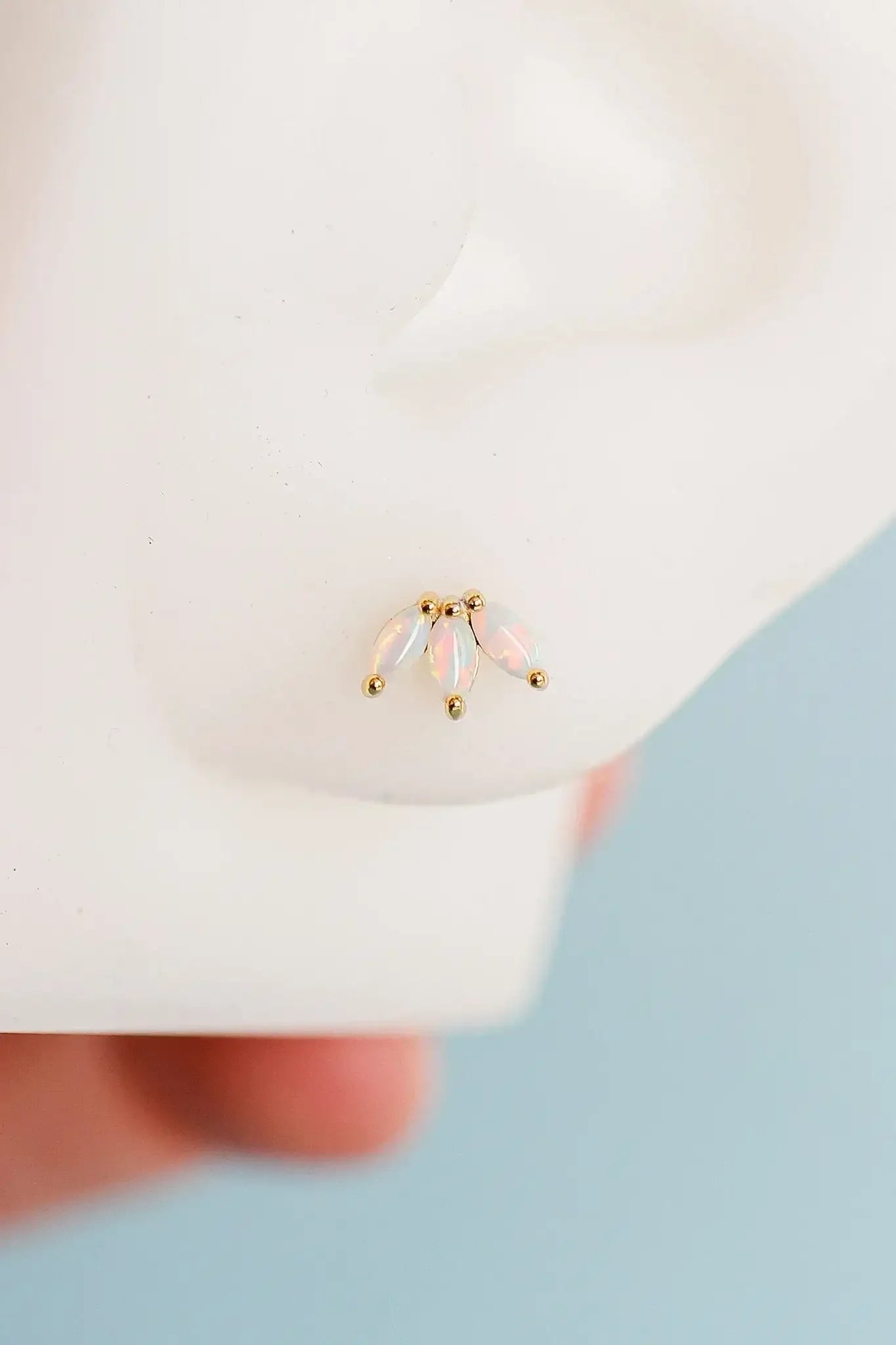 A small gold earring featuring three opalescent, leaf-shaped stones is displayed on a mannequin ear, with a blurred hand in the background, suggesting a setting focused on jewelry presentation.