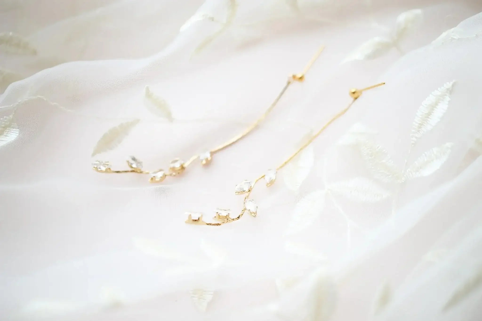 Delicate gold earrings, designed in a floral vine pattern with sparkling gemstones, rest atop soft, sheer fabric embroidered with leaf motifs, creating an elegant and romantic display.