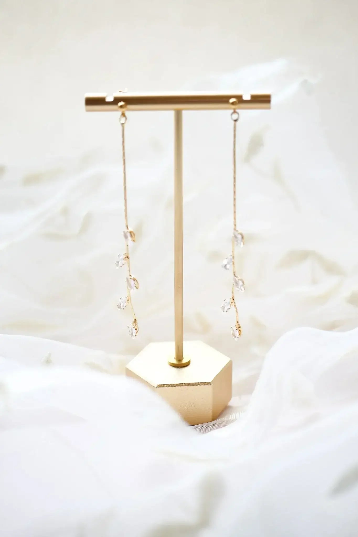 A pair of elegant, dangling earrings with crystal-like embellishments hang from a minimalist gold display stand, set against a soft, flowing white fabric background.