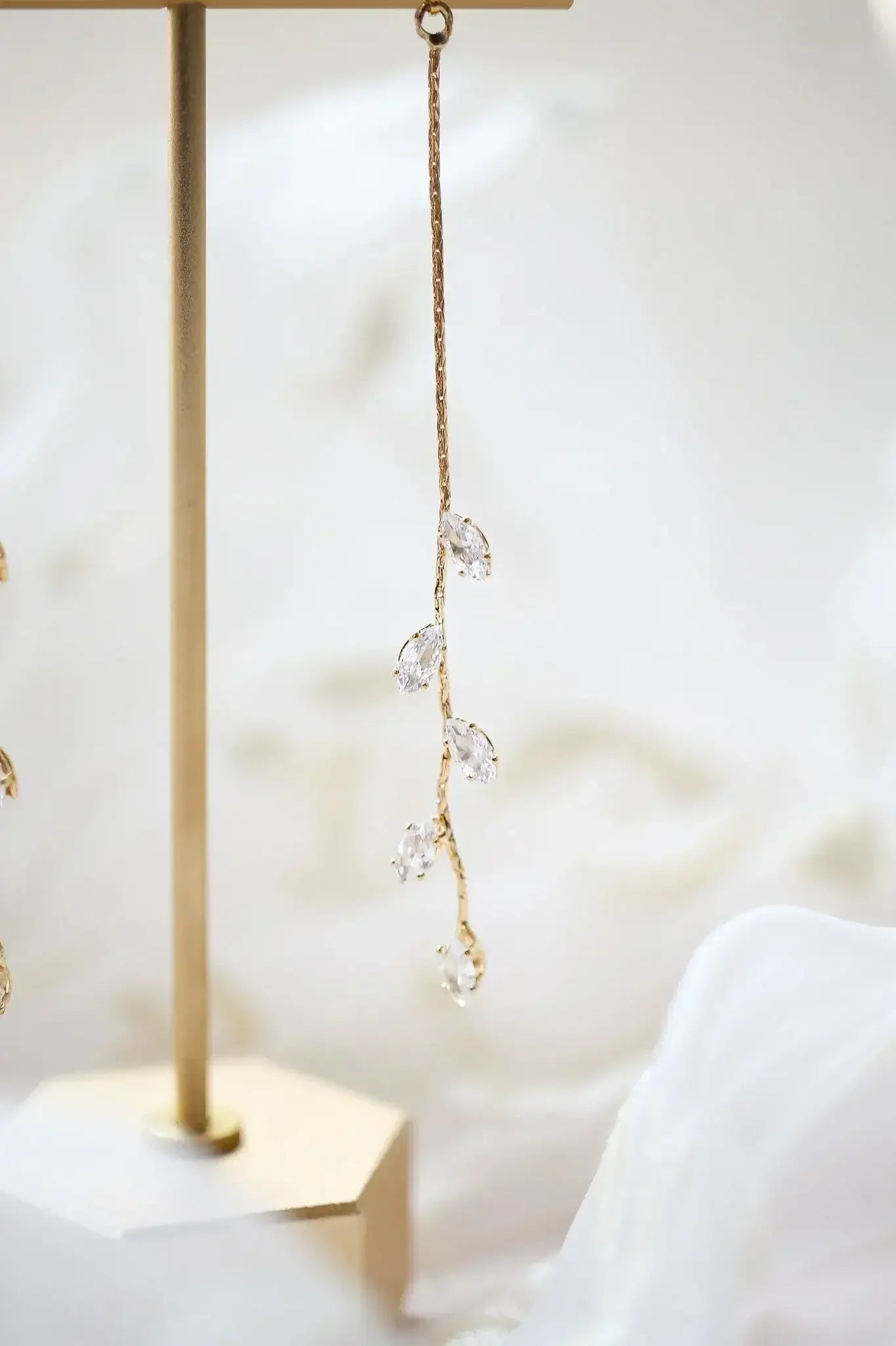 A delicate gold earring hangs from a stand, featuring a twisting design adorned with clear gemstones. The soft, blurred background of fabric adds an elegant touch to the display.