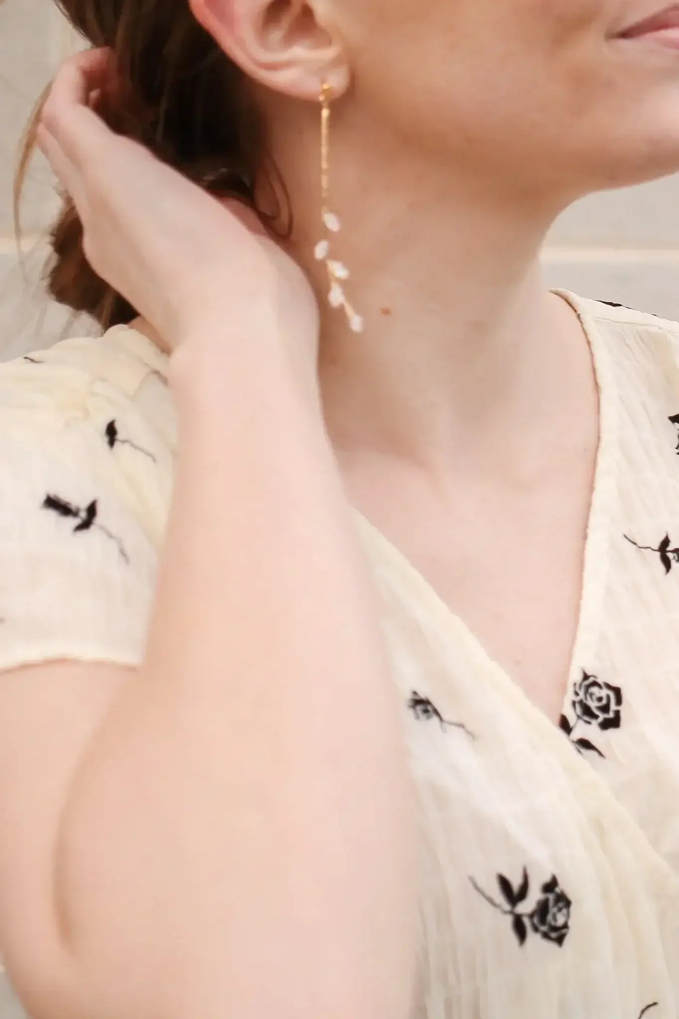 A woman wears a detailed, long gold earring with floral accents, gently touching her hair. She is dressed in a light, patterned blouse against a softly blurred neutral background.