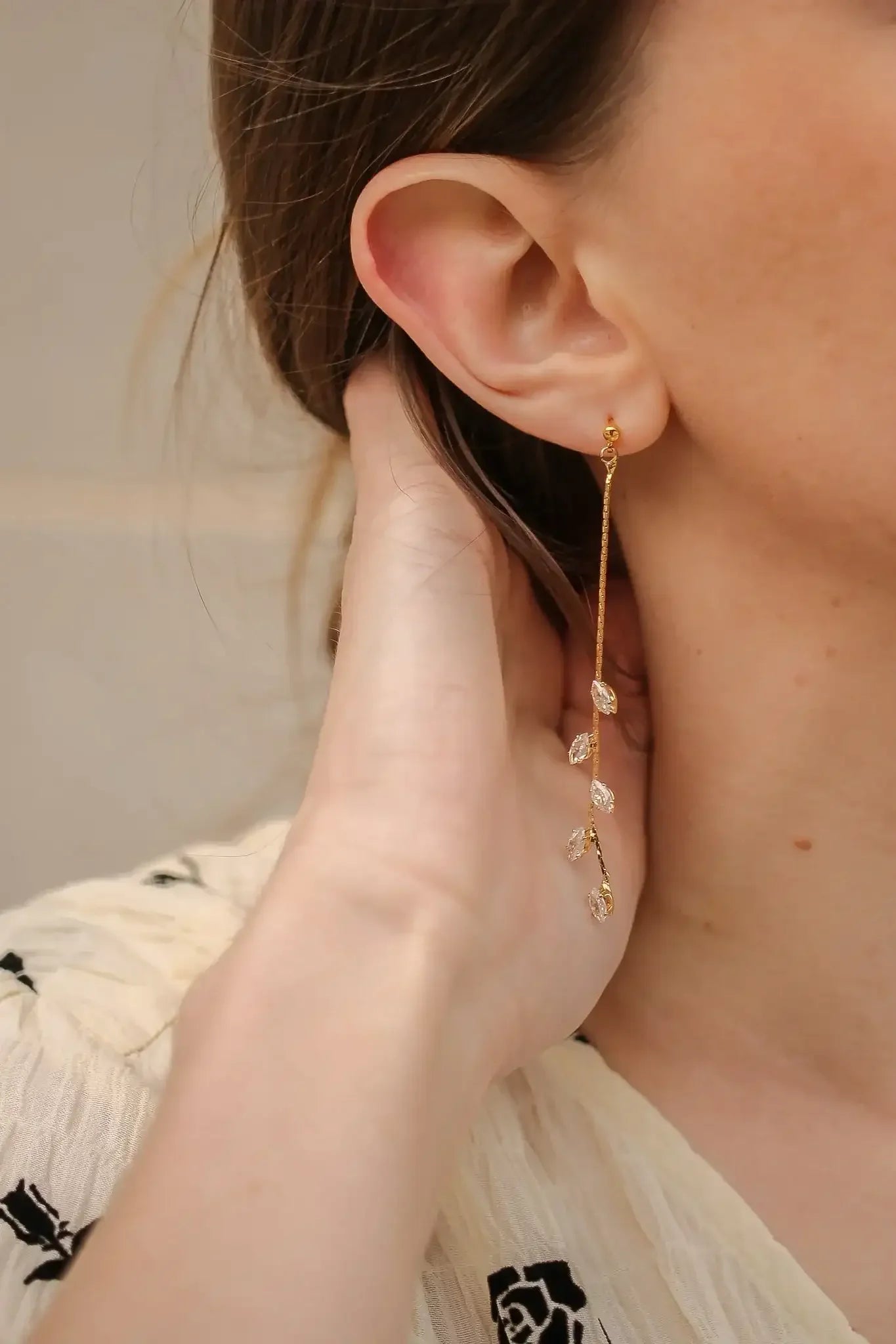 A hand gently touches the neck near an ear adorned with a delicate gold earring featuring crystal leaf designs, set against a soft, light-colored textured background.